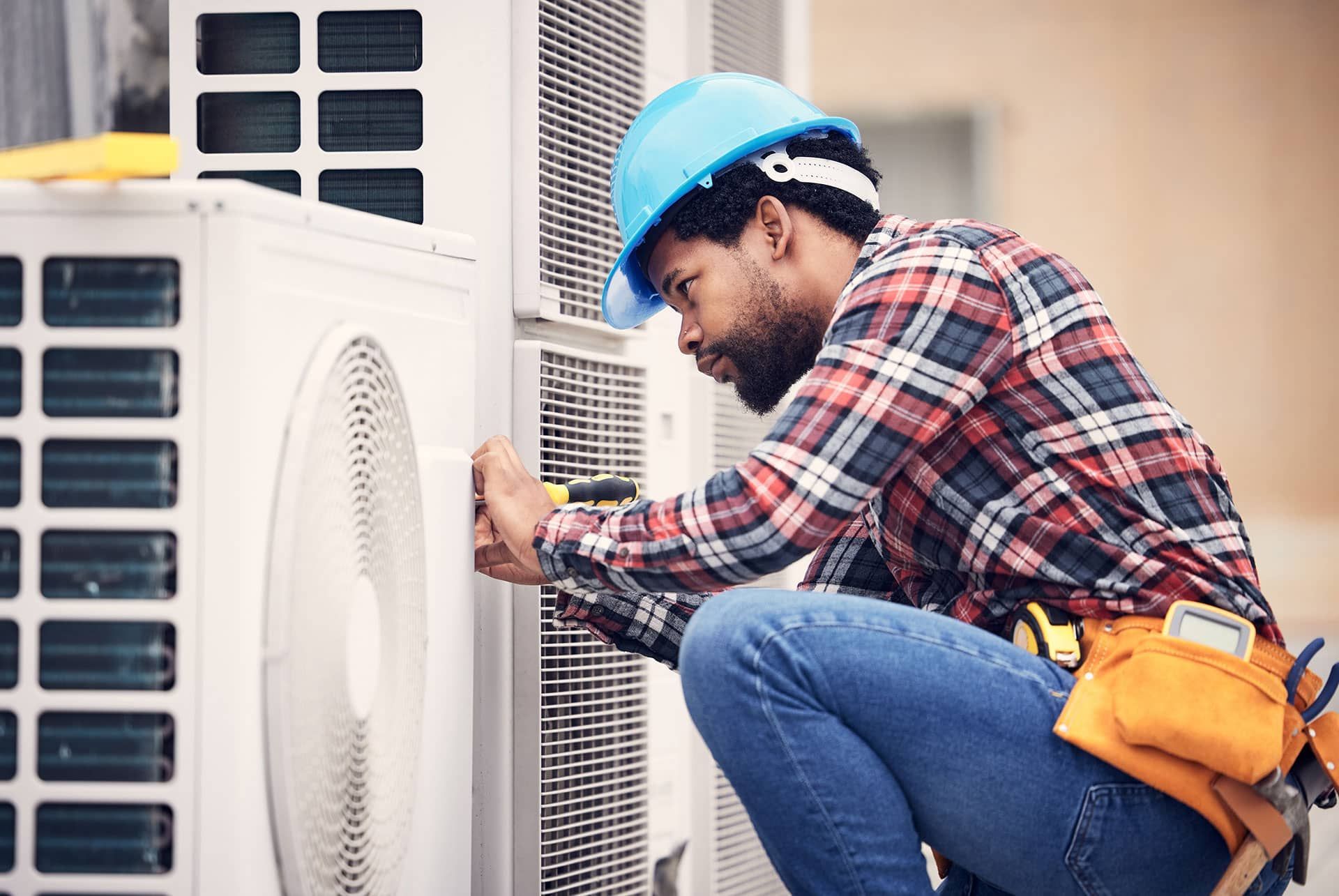 HVAC technician in blue hard hat works on an air conditioning unit outside.
