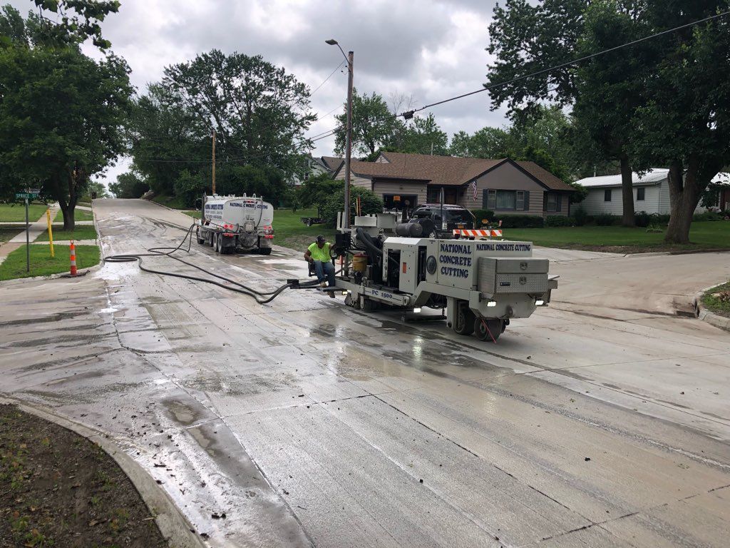 Road construction on a residential street. A machine grinds pavement. Trucks and workers are present.