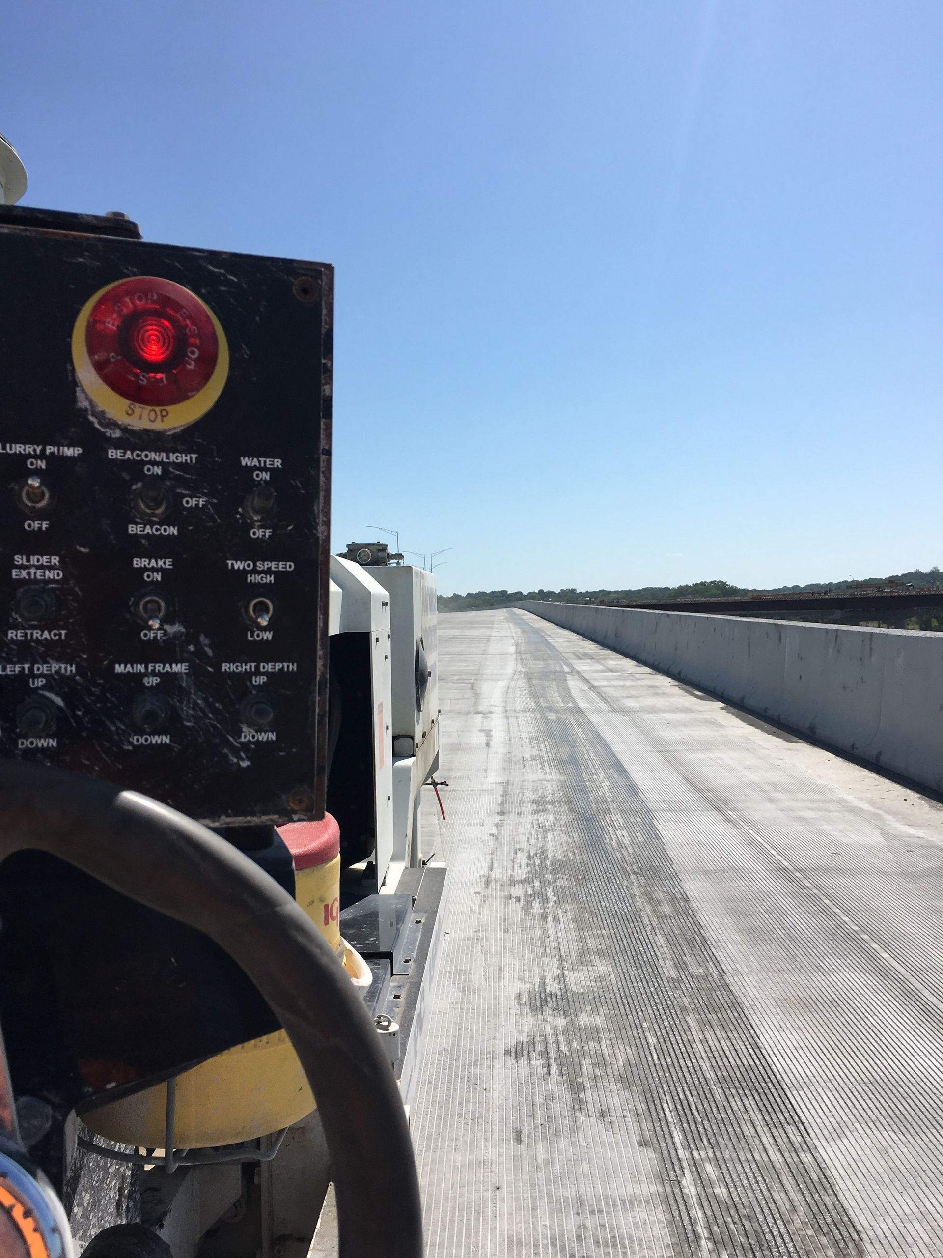 View from a vehicle's dashboard, showing a freshly textured concrete bridge under a clear, blue sky.