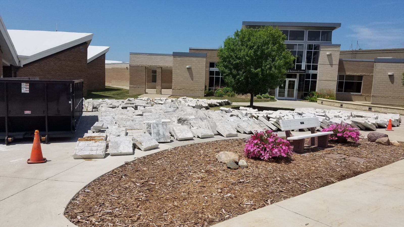 Gray stone slabs piled on a concrete area outside a light brown building with a tree and a bench.