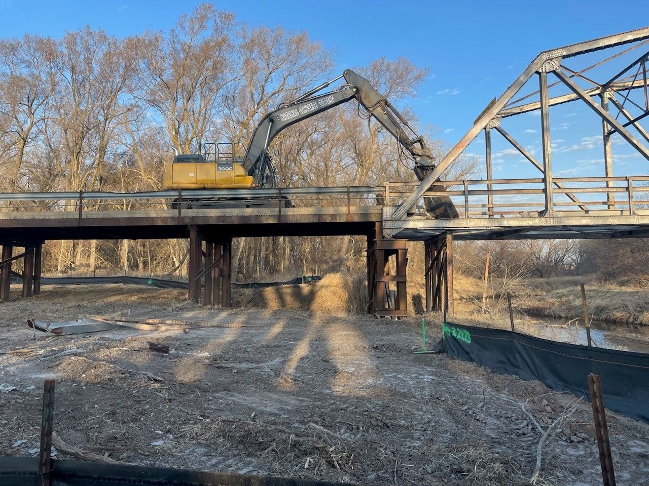An excavator demolishes an old metal bridge in a wooded area on a sunny day.
