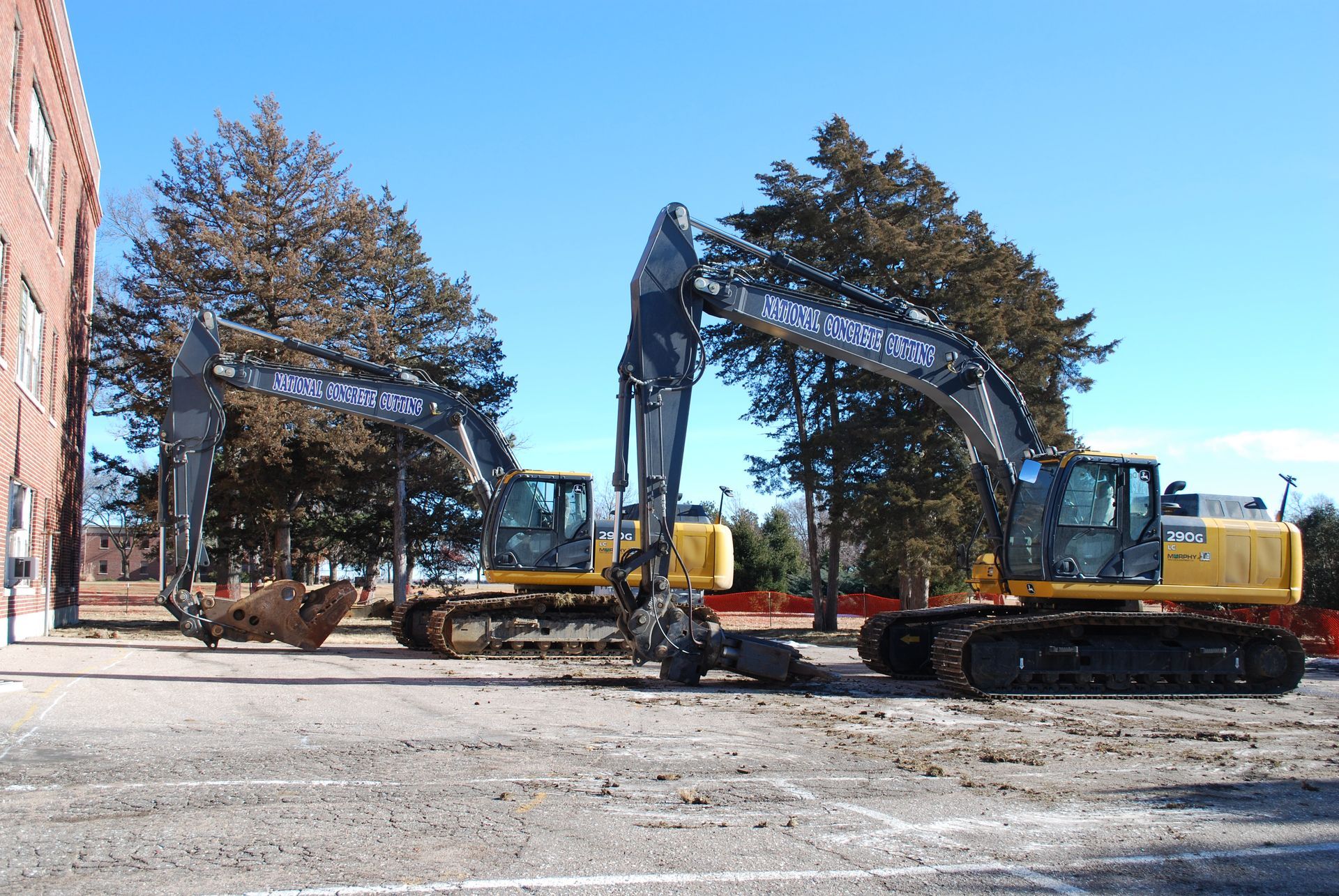 Two yellow excavators parked on a concrete lot next to a brick building, under a blue sky.