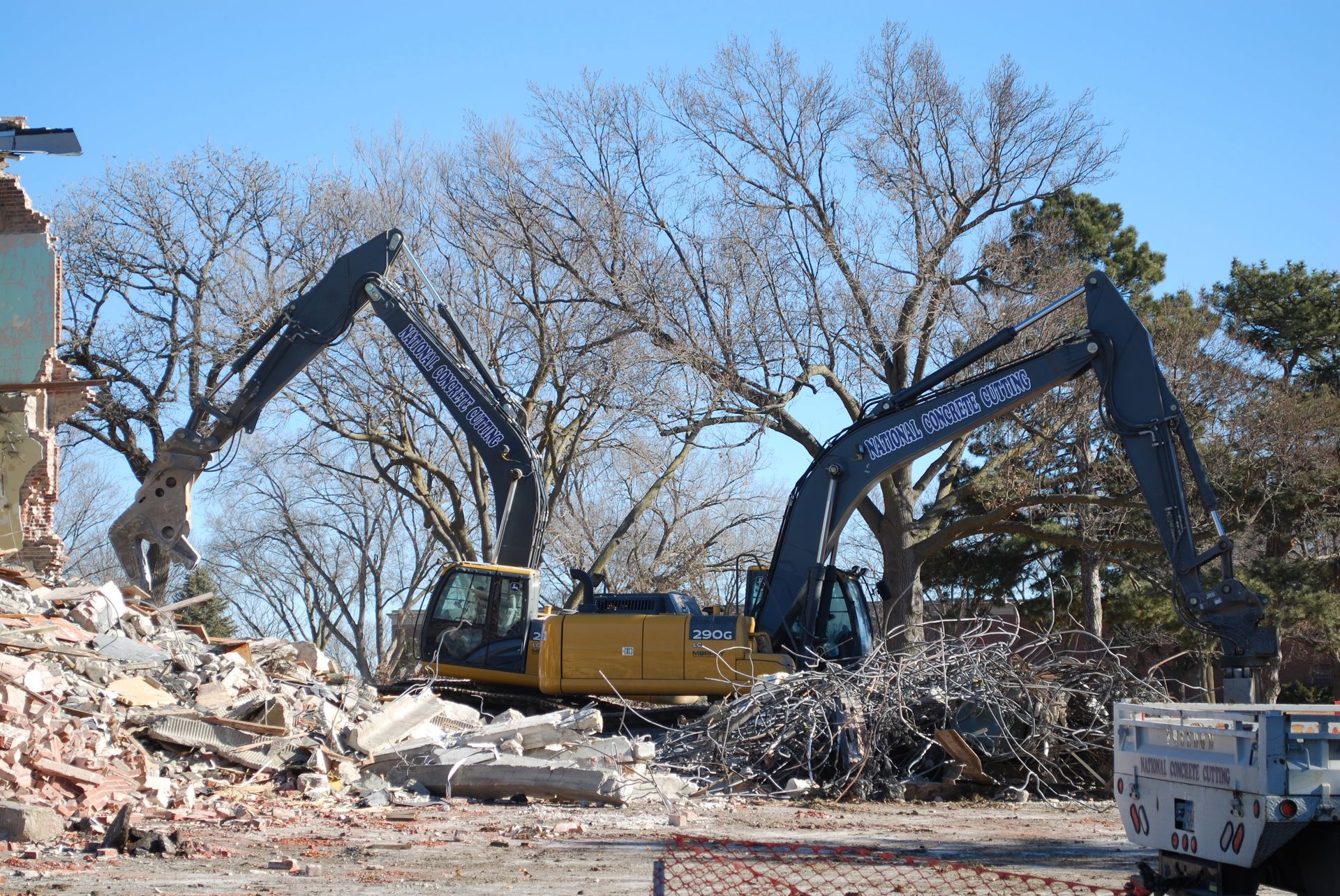 Two excavators demolish a building under a sunny sky. Debris and bare trees are visible.