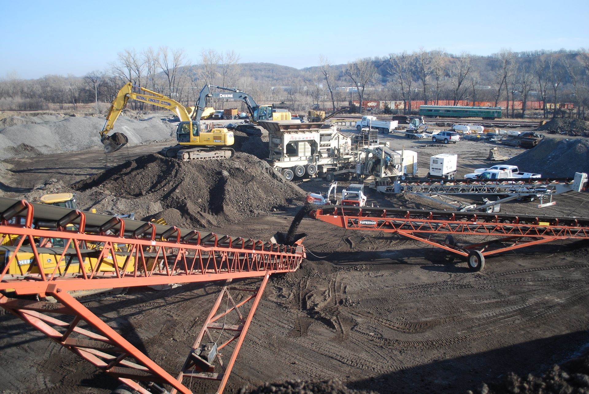 Construction site with excavator and conveyor belt, mountains in background.