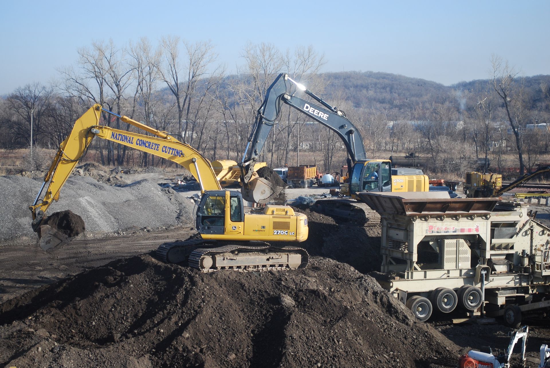 Two excavators working on a construction site. One yellow, one blue, moving earth.