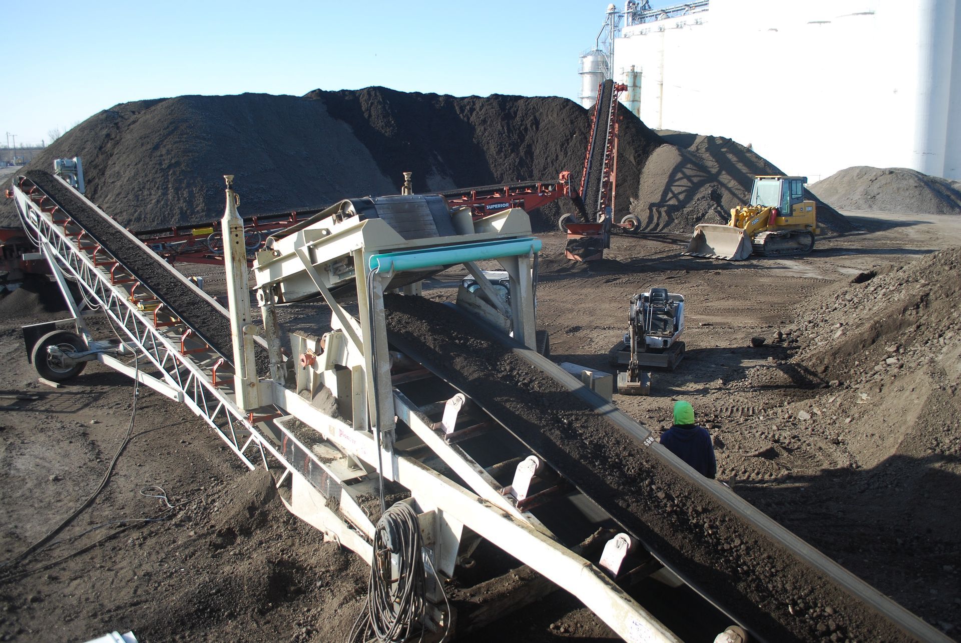 Conveyor belt depositing dark material onto a pile. Construction site with machinery and a silo in background.