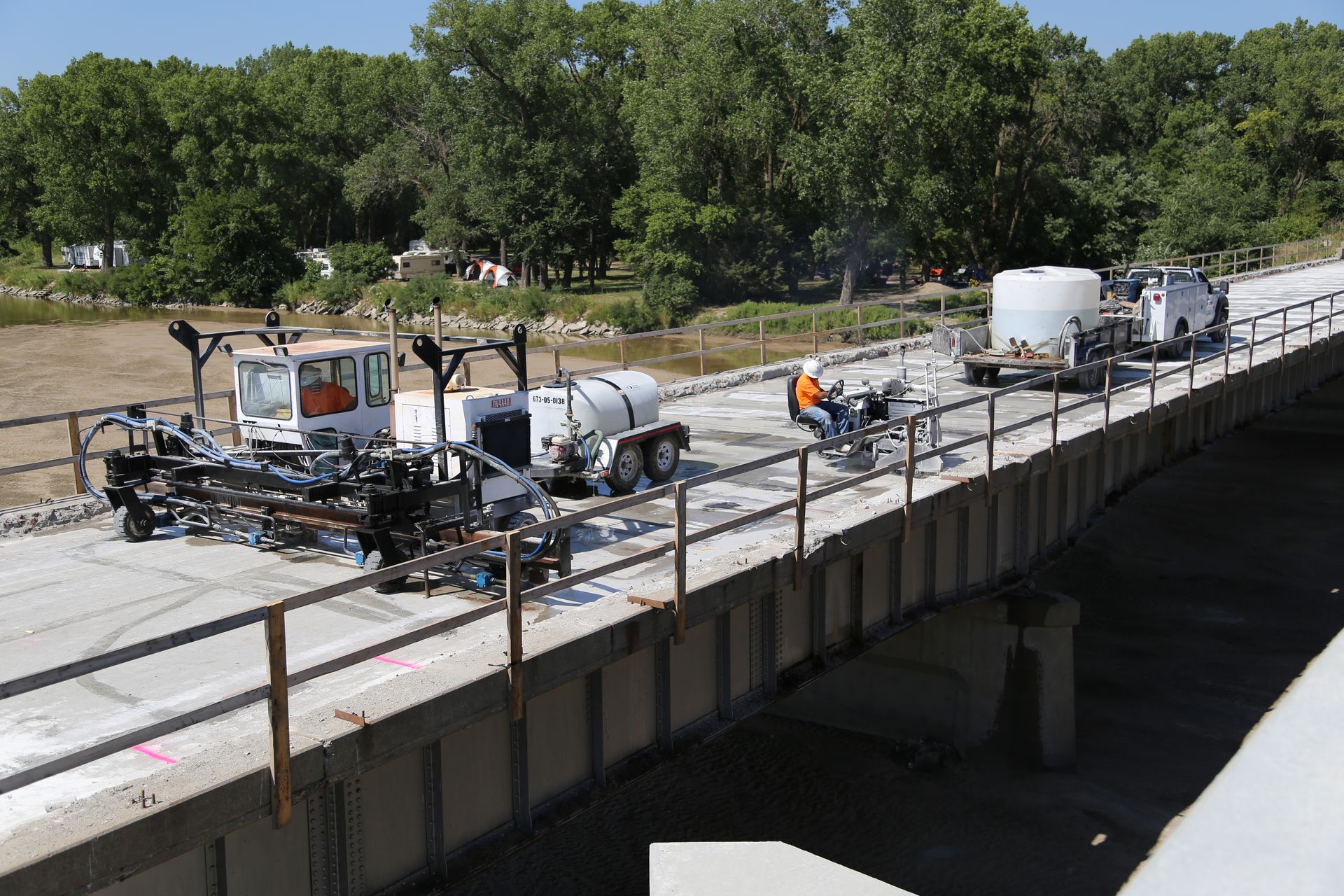 Bridge repair: machinery and workers on a concrete bridge deck, trees and water in background.