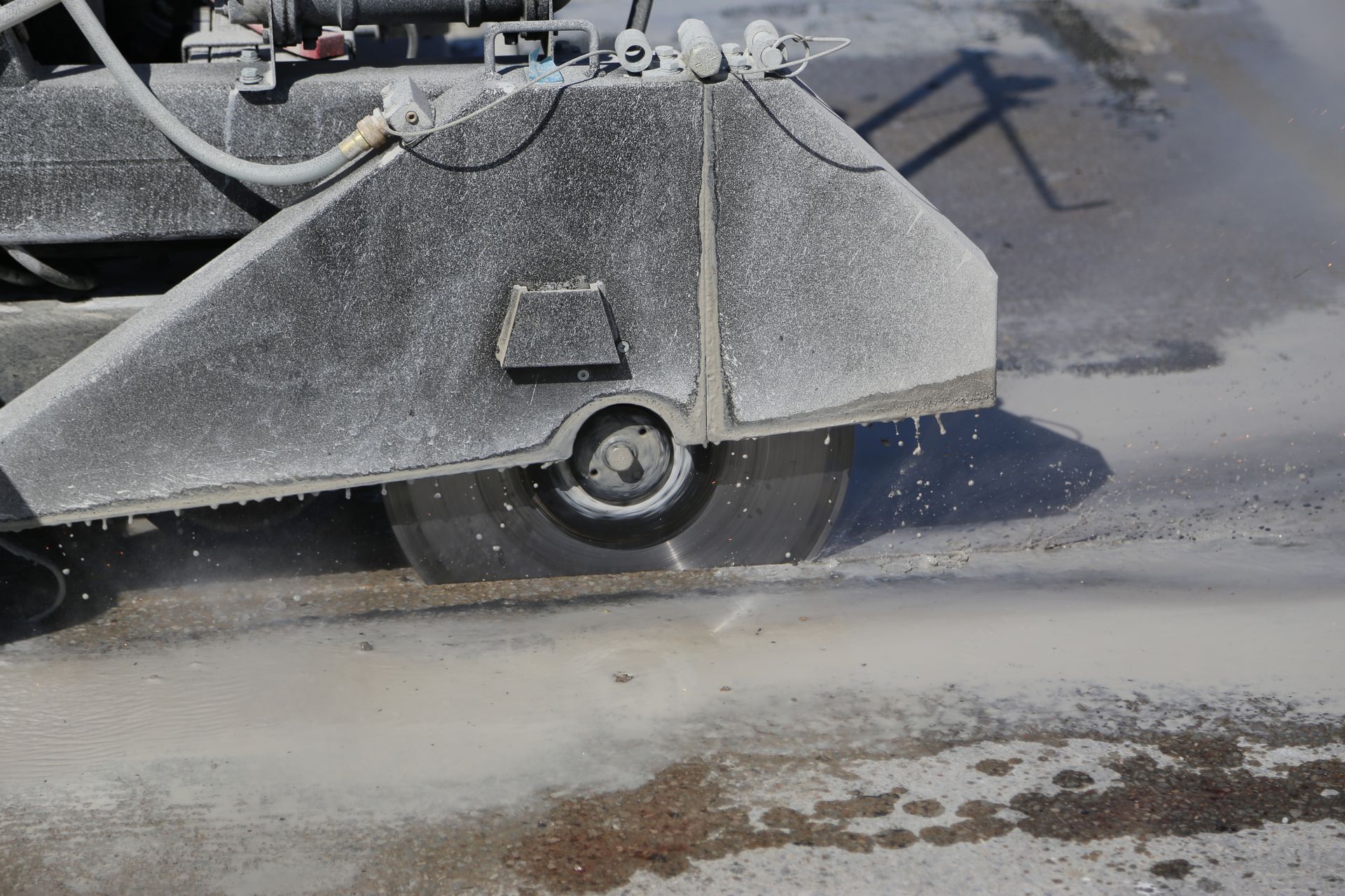 Concrete saw cutting pavement, water spraying from blade, dusty gray setting.