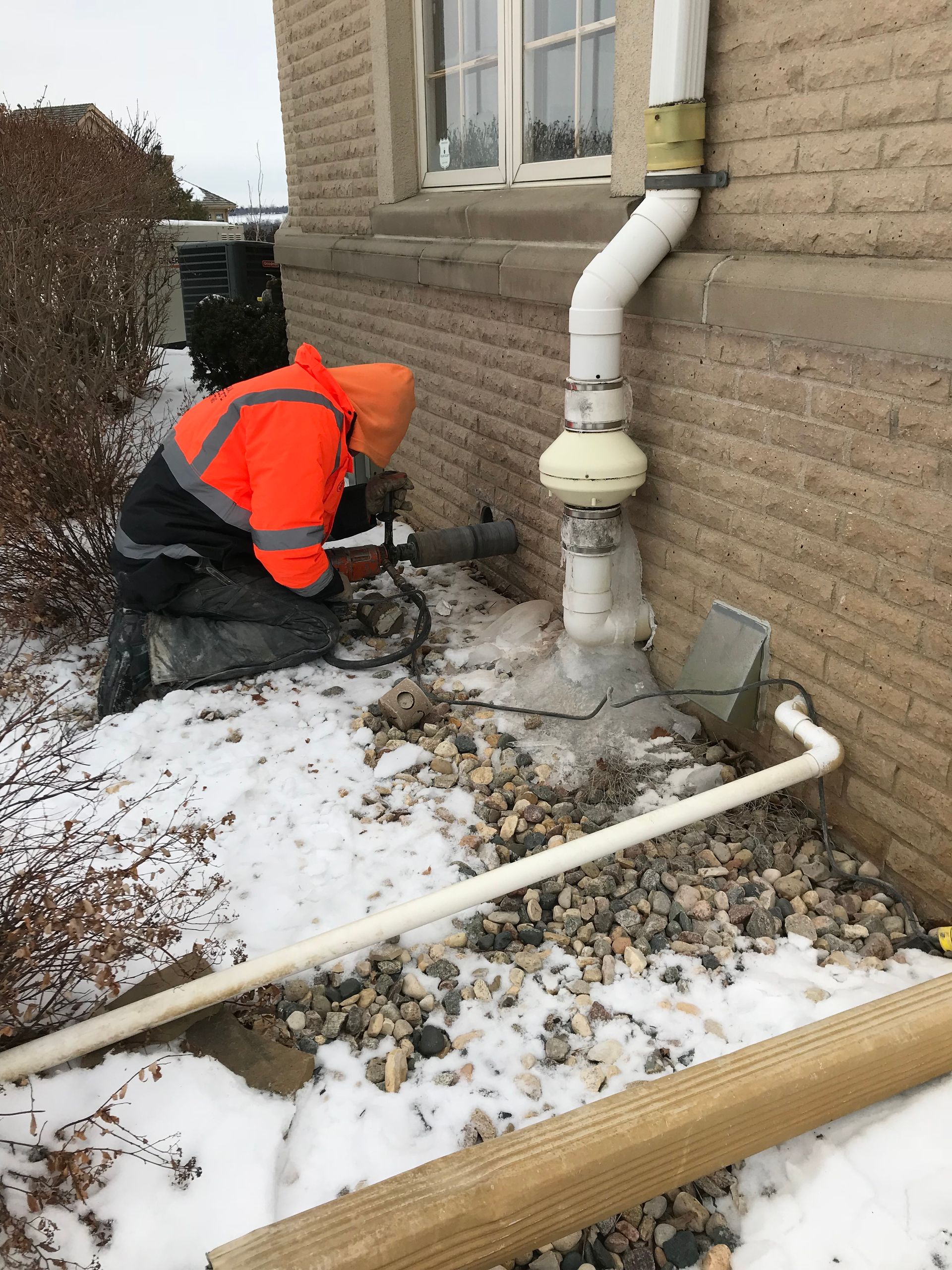 Person in orange workwear working near a building's foundation and drainage pipe in a snowy setting.