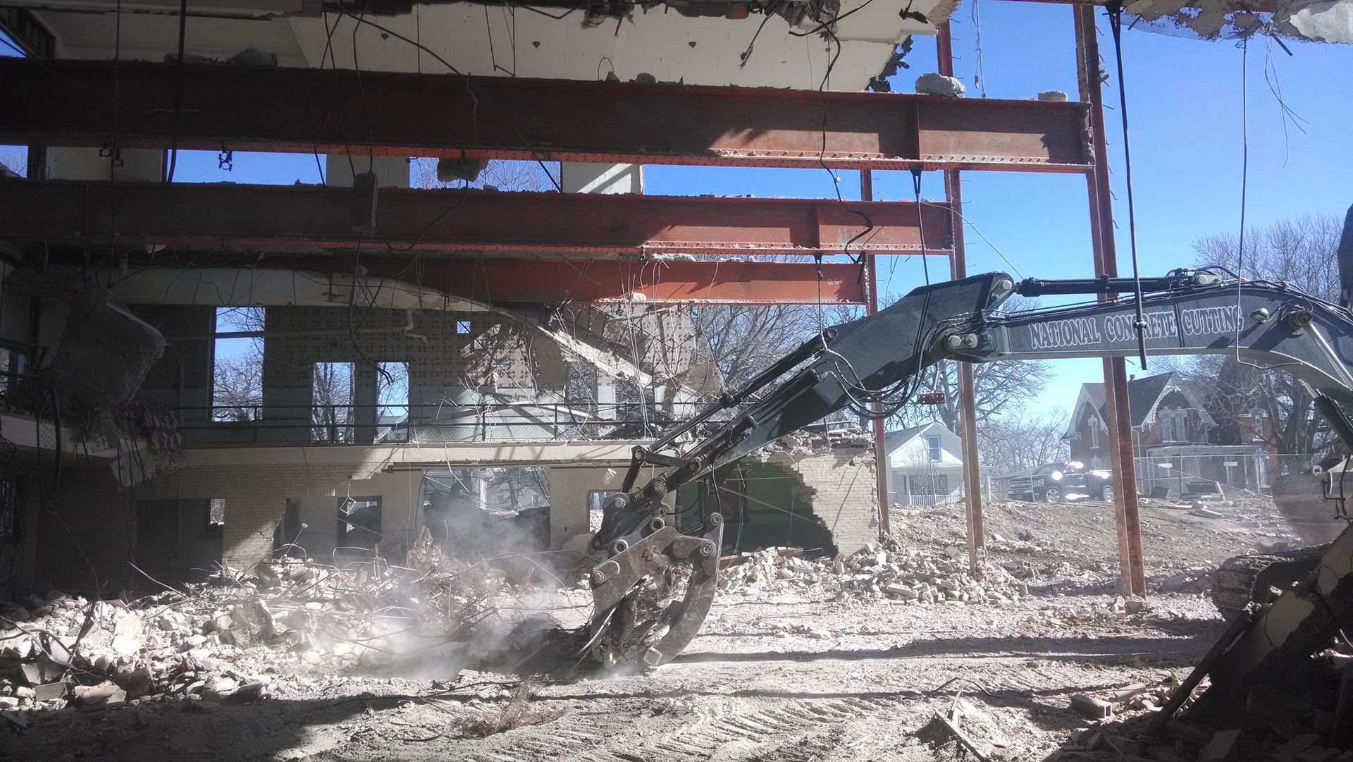 Excavator demolishing a building; debris and dust in the air. Steel beams and blue sky visible.