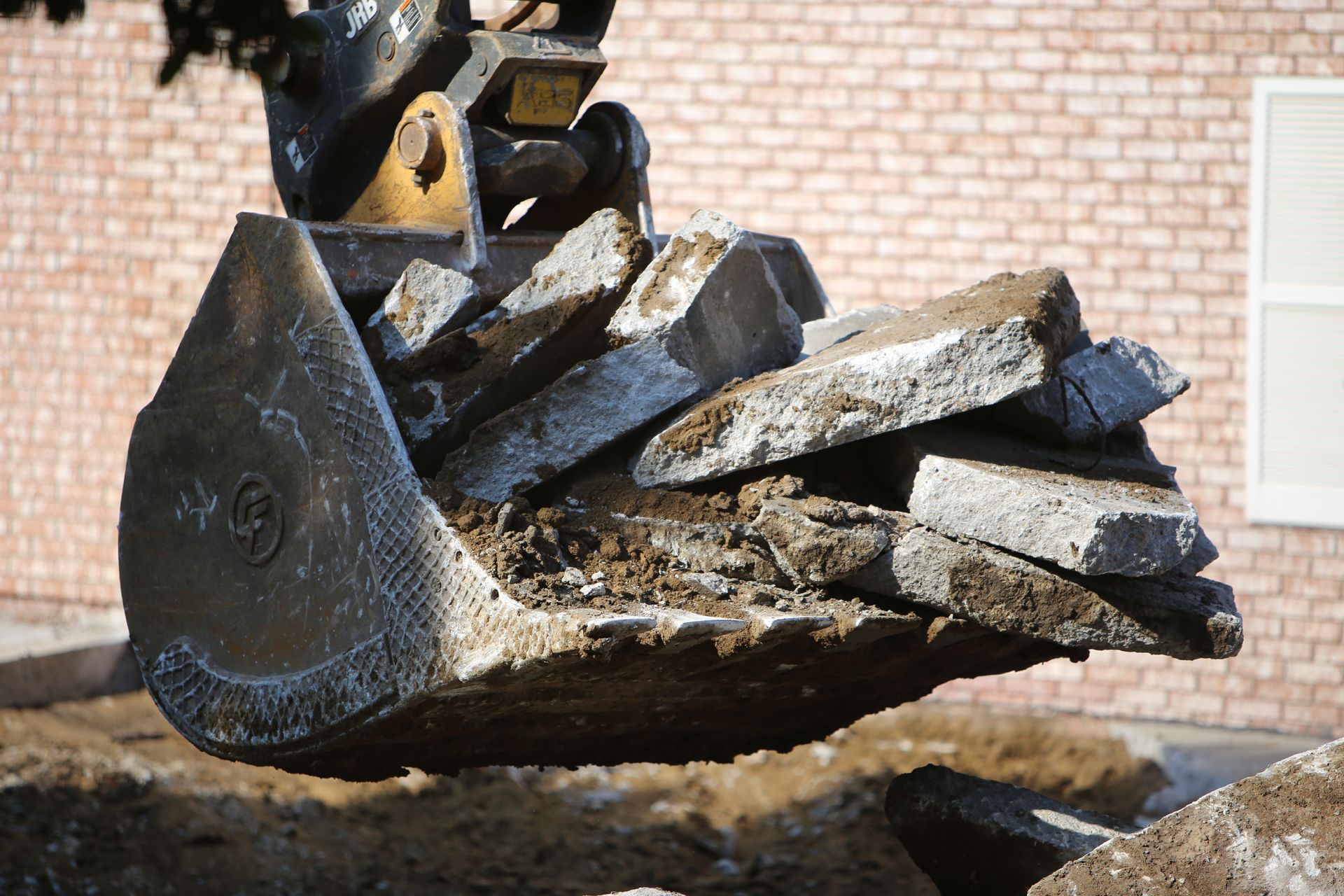 Excavator bucket holding broken concrete slabs against a brick building.