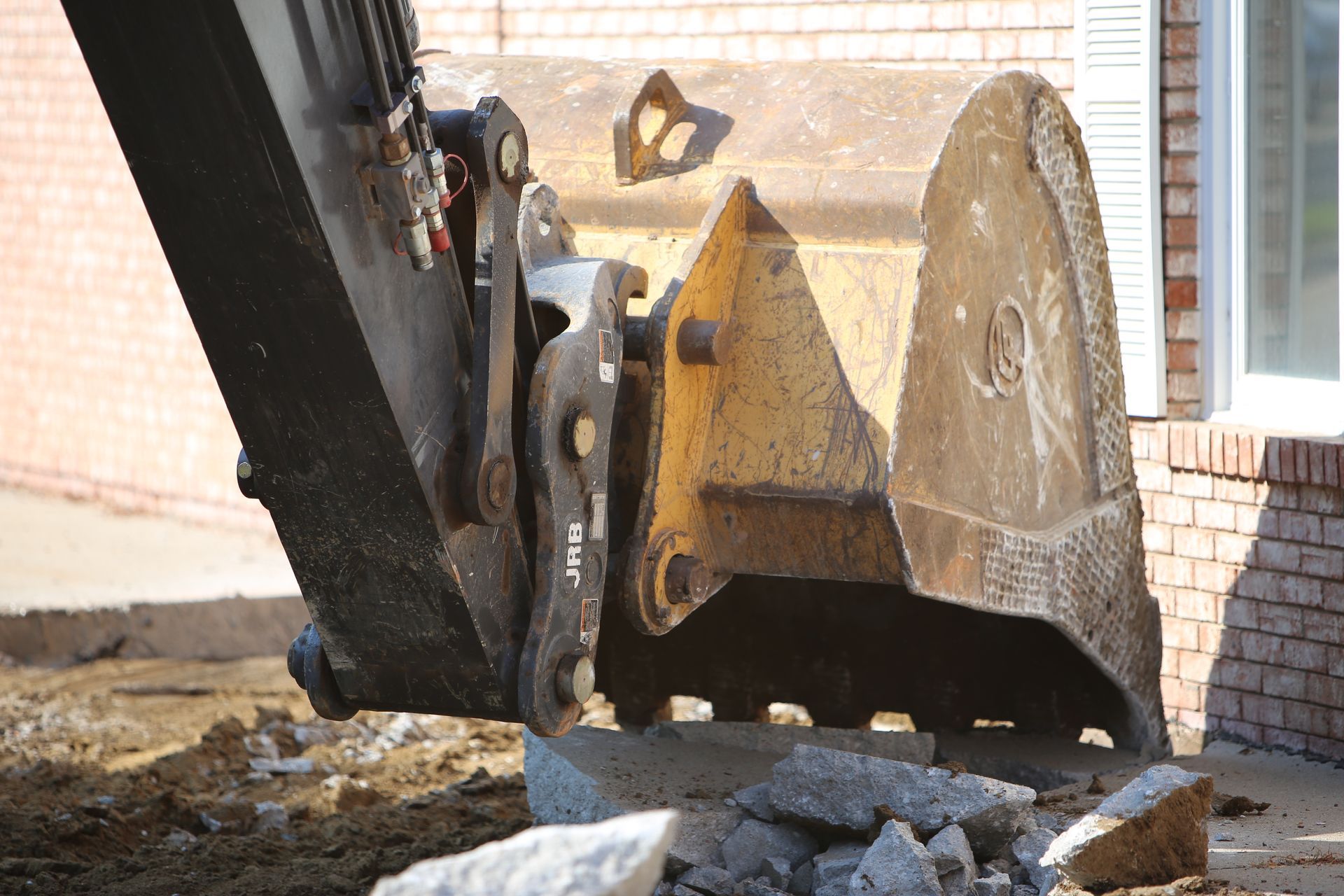Excavator bucket breaking up concrete near a brick building with a white shuttered window.