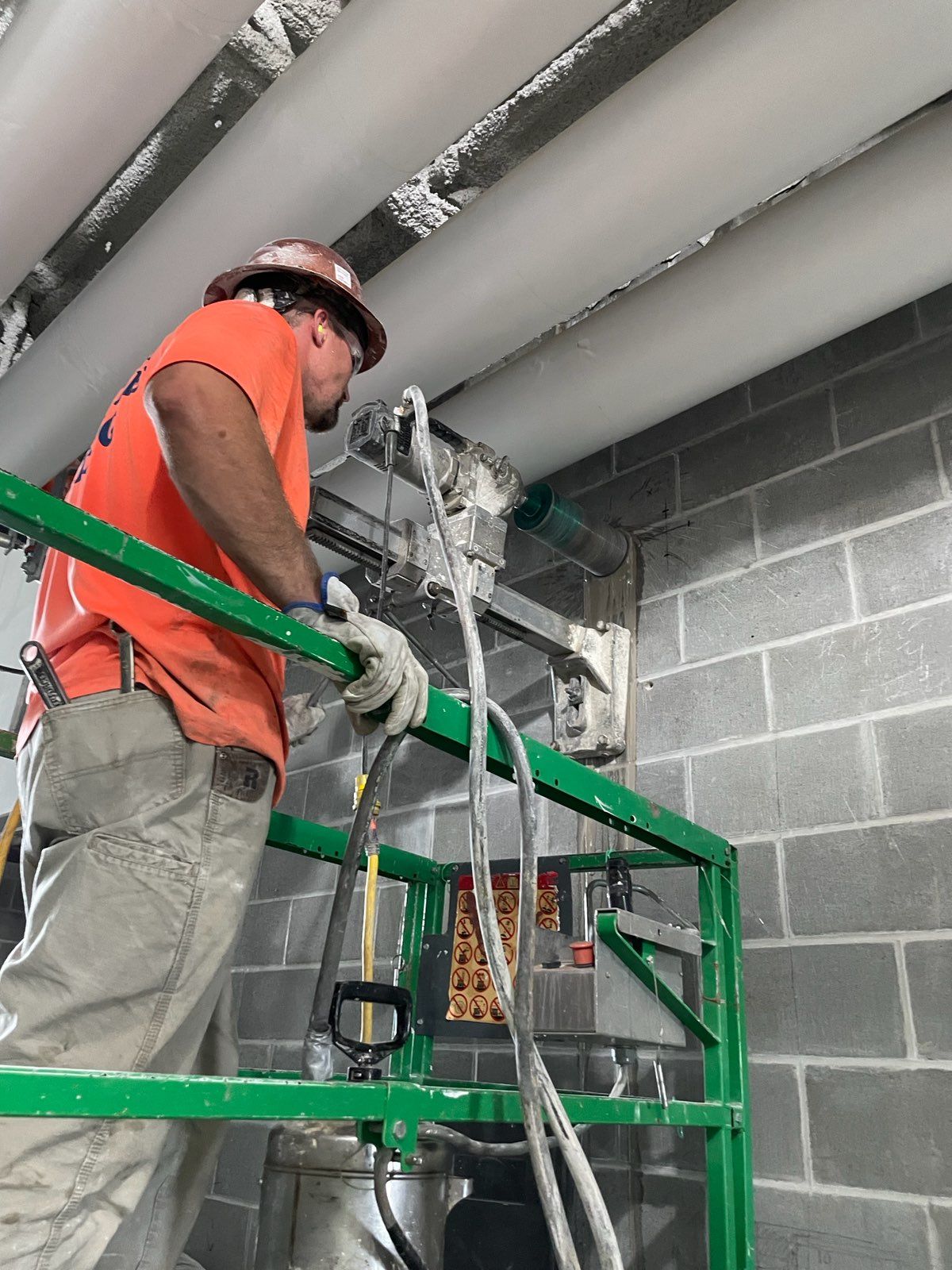 Construction worker cutting concrete block wall while on a green lift. He wears an orange shirt and hard hat.