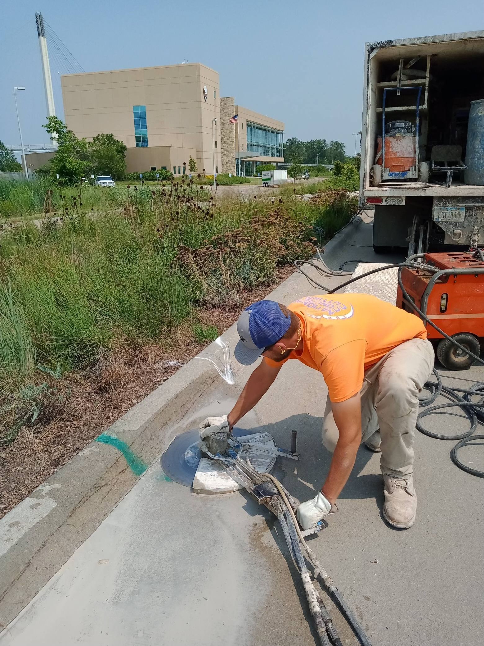 Man in orange shirt grinding concrete curb. Truck with equipment in background, sunny day.