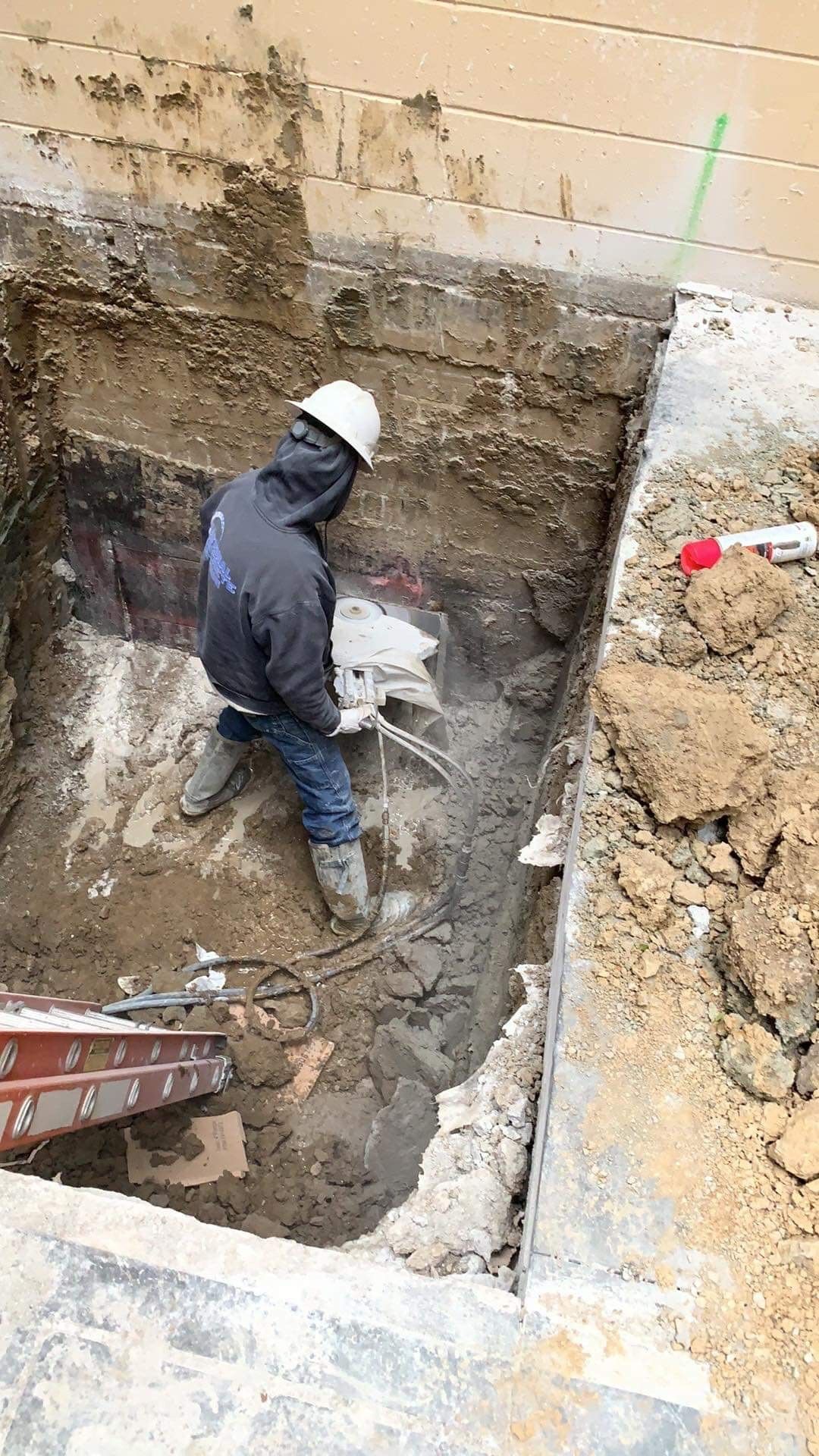 Person in a hard hat uses a jackhammer in an excavated area. Dirt and debris surround the concrete structure.