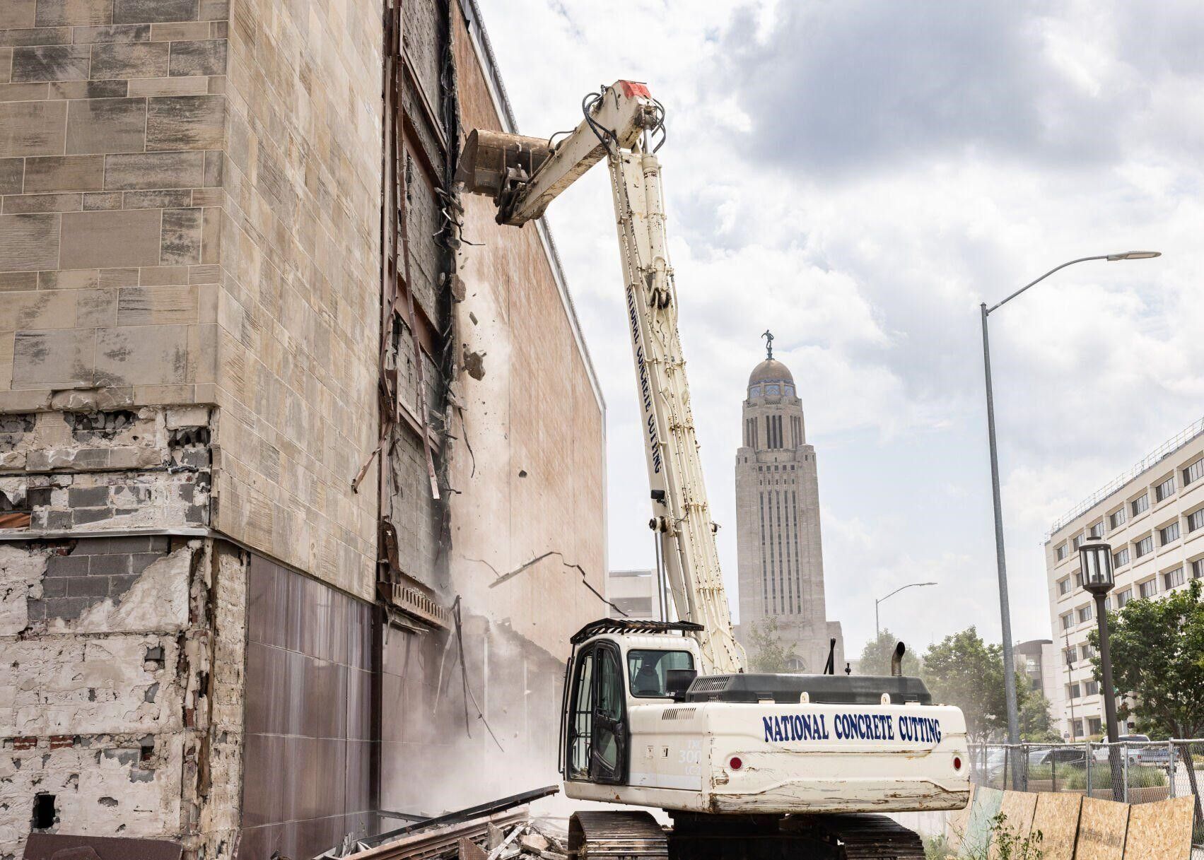 A long-reach excavator demolishes a building wall, with the Nebraska State Capitol in the background.