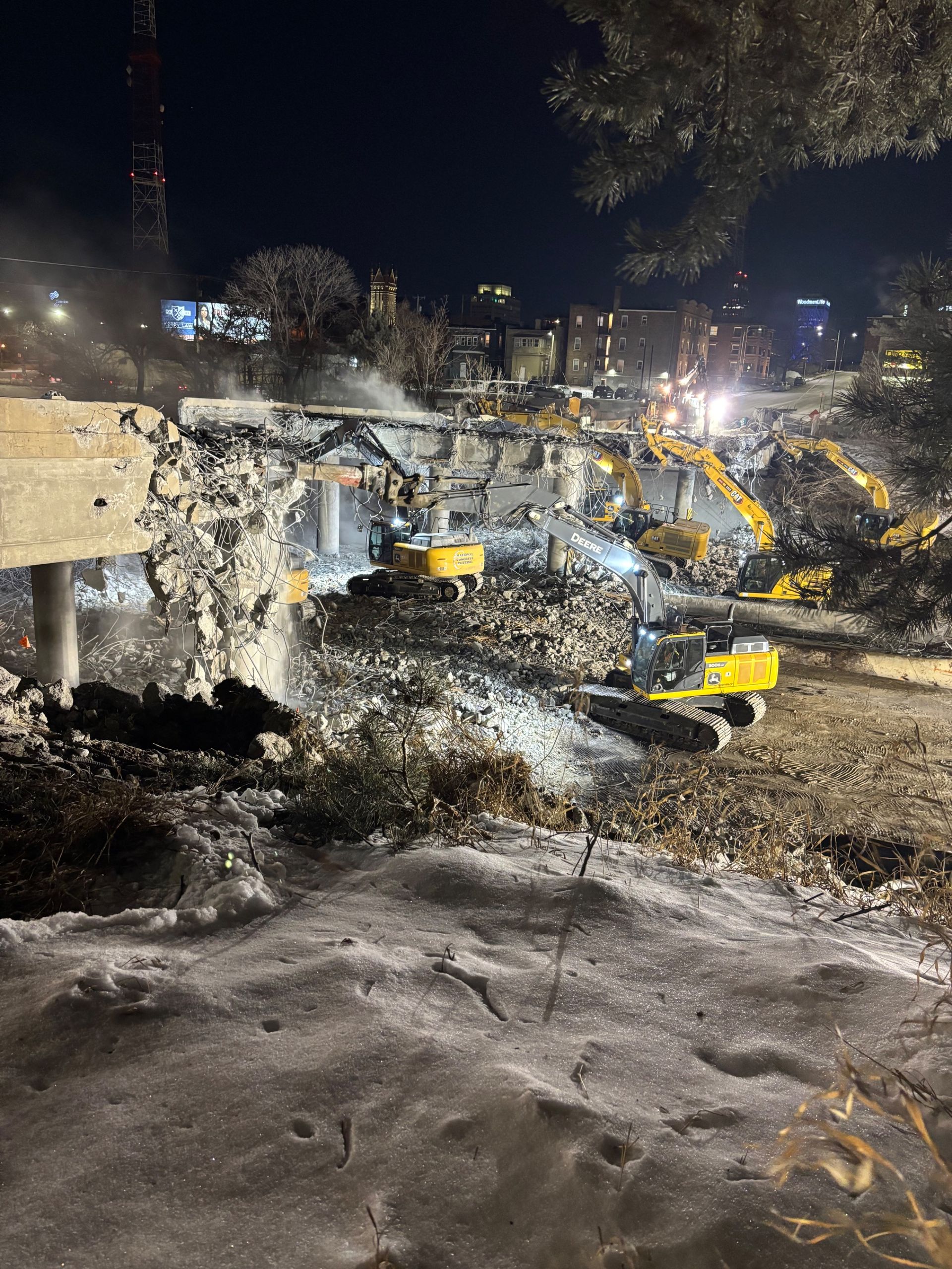 A yellow John Deere excavator actively tears down the structure, producing dust against a clear blue sky. A yellow John Deere excavator actively tears down the structure, producing dust against a clear blue sky.