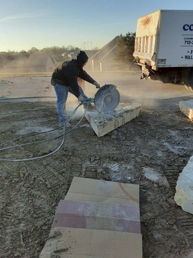 Person cutting stone with a circular saw outdoors, creating dust. Truck in background.