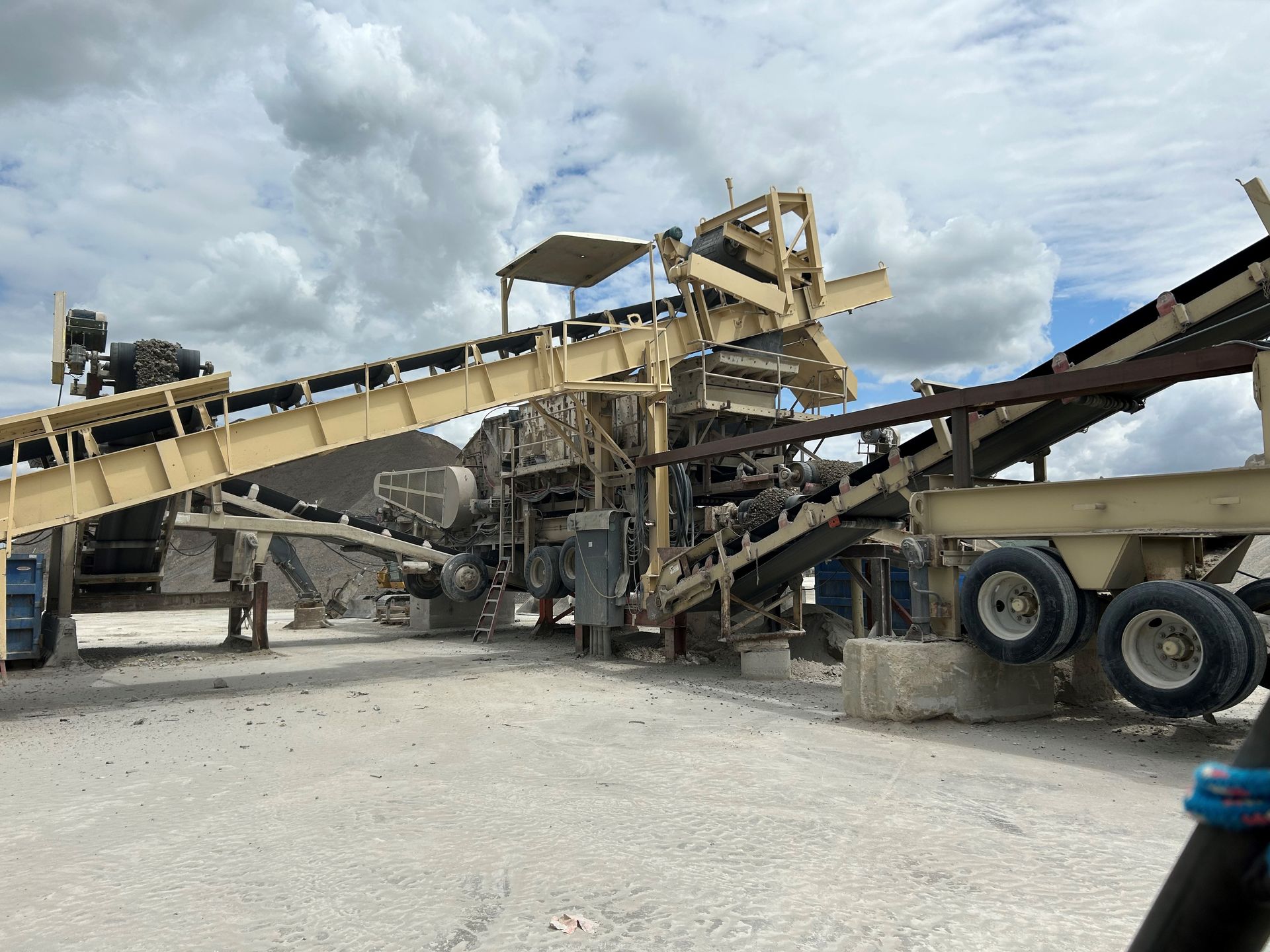 A beige rock crusher with conveyor belts operates outdoors on a cloudy day.