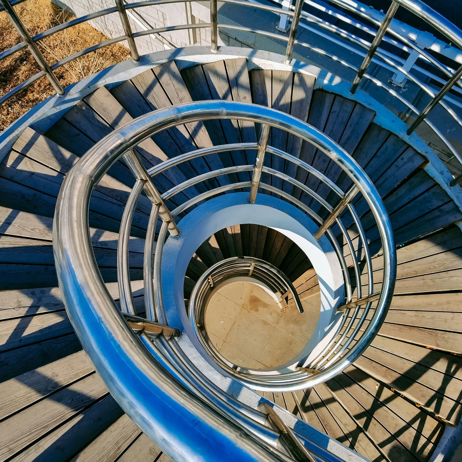 Spiral metal staircase with blue railings viewed from above, curving down to a sandy landing below