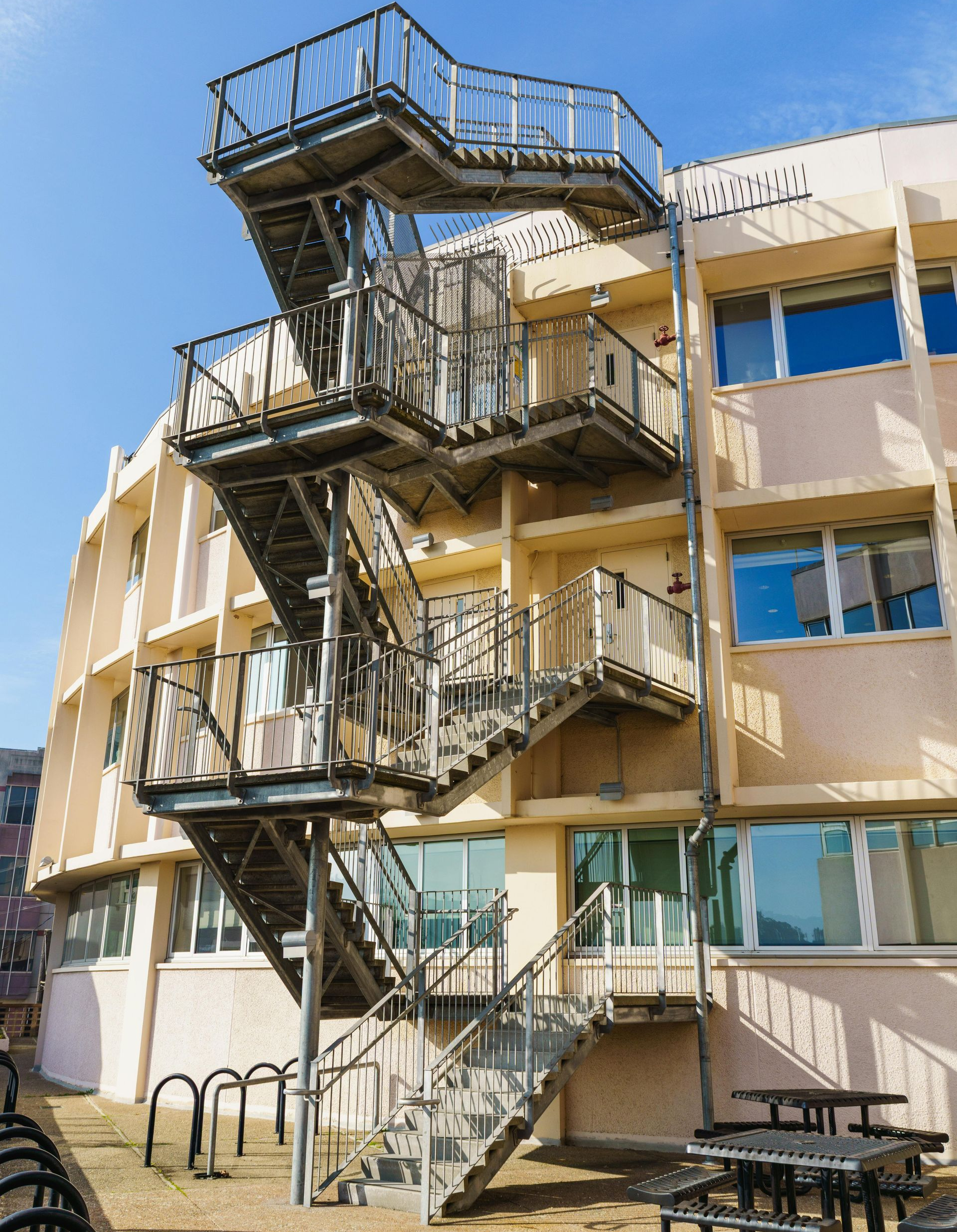 An exterior view of a multi-story, beige building with a prominent, detached metal fire escape staircase on the side.