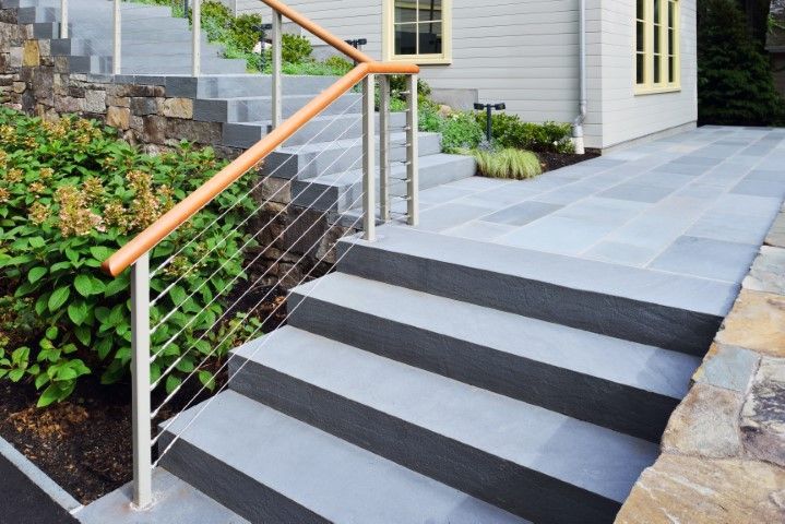 Stone steps and walkway leading to a gray house, with a wooden handrail and greenery.