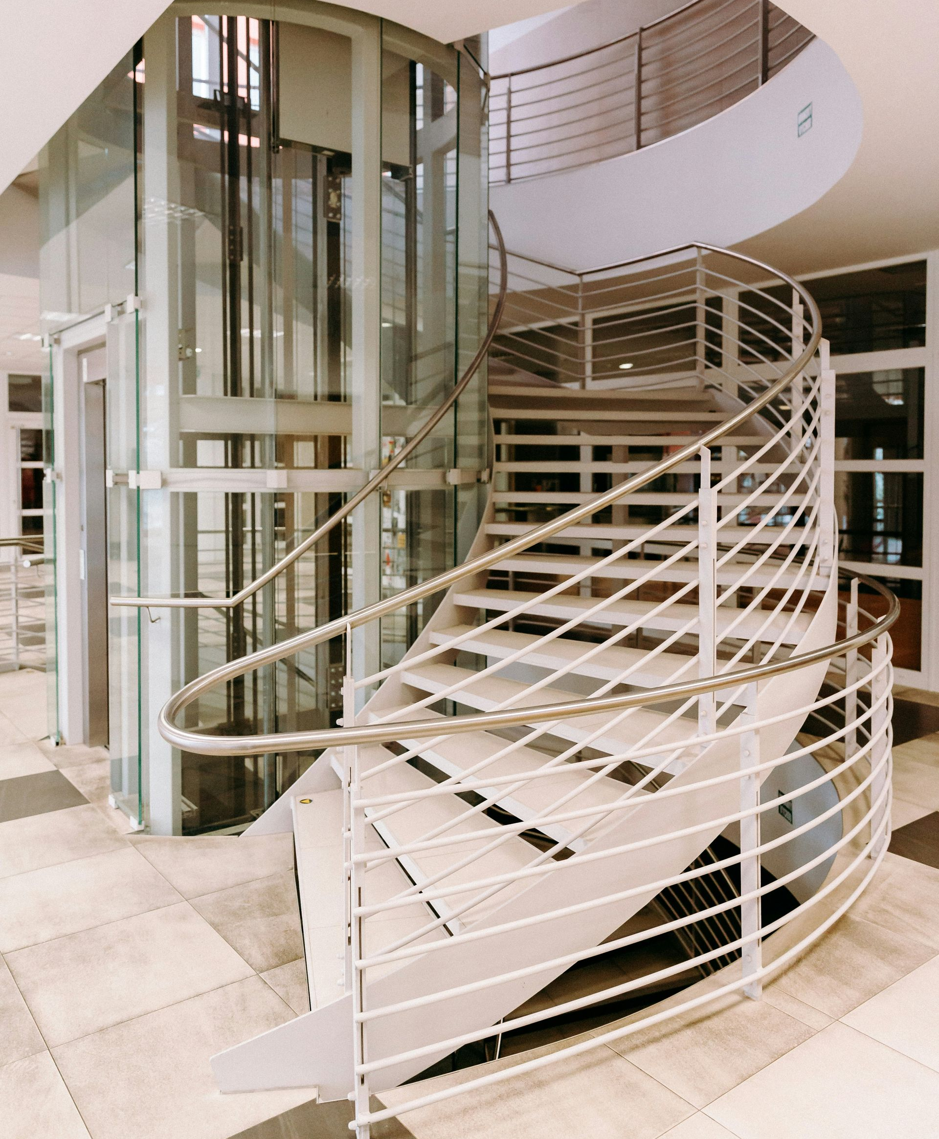 A white spiral staircase winding around a cylindrical glass elevator in a bright, modern building lobby.