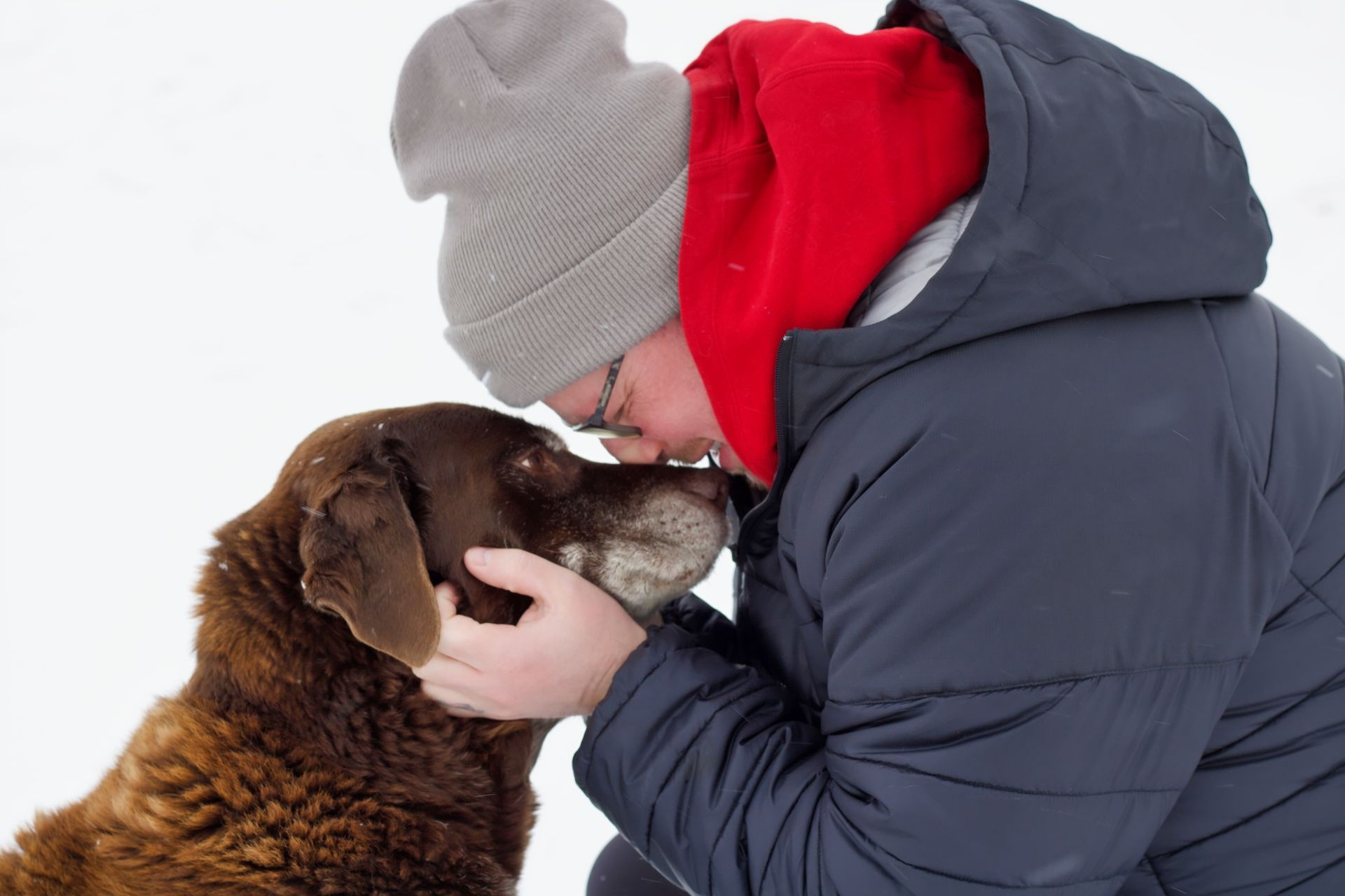 Person in winter coat and hat nuzzles a brown dog's face in the snow.
