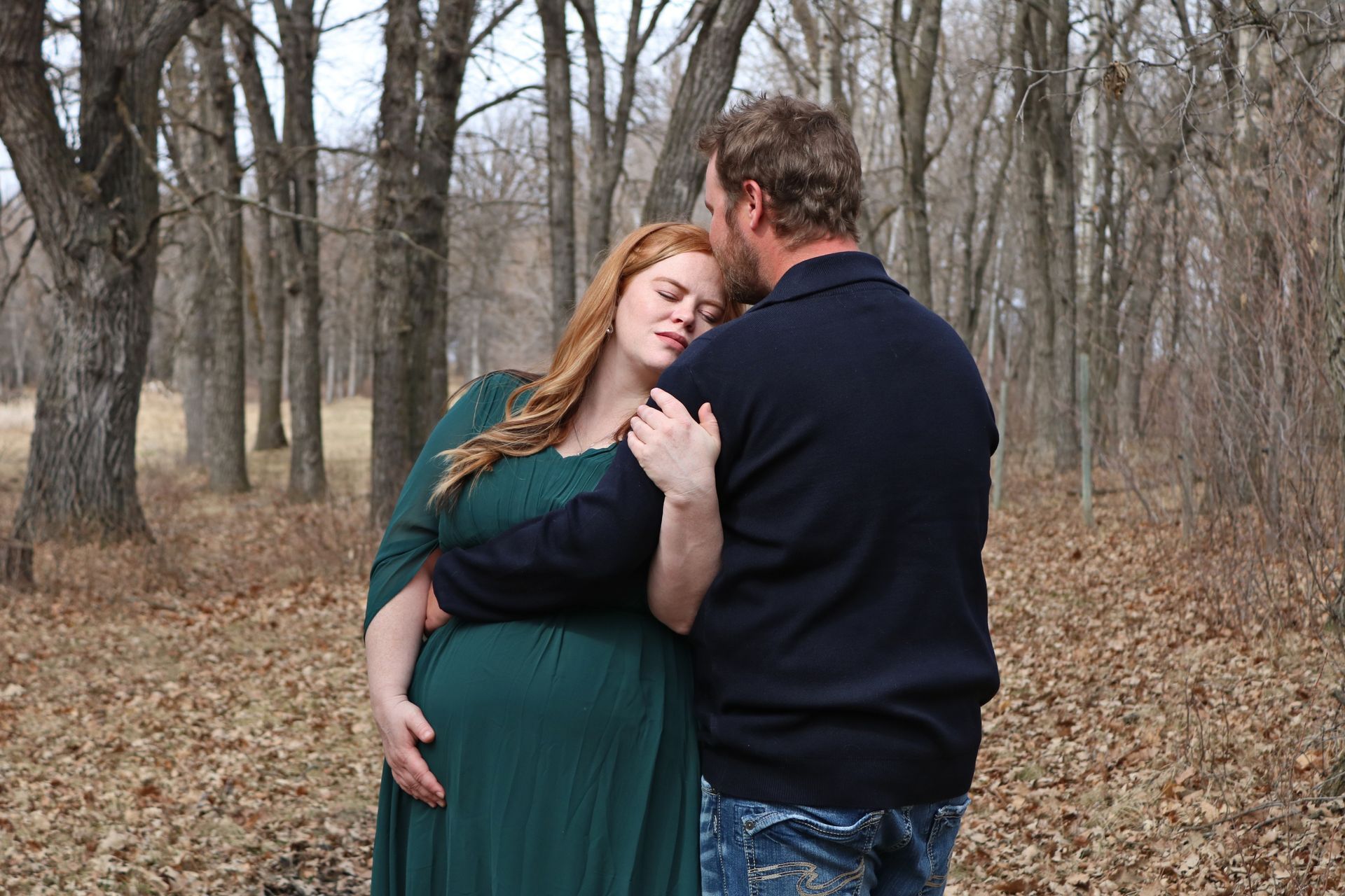 Couple embracing in a leafless wooded park, woman in green dress and man in dark sweater.