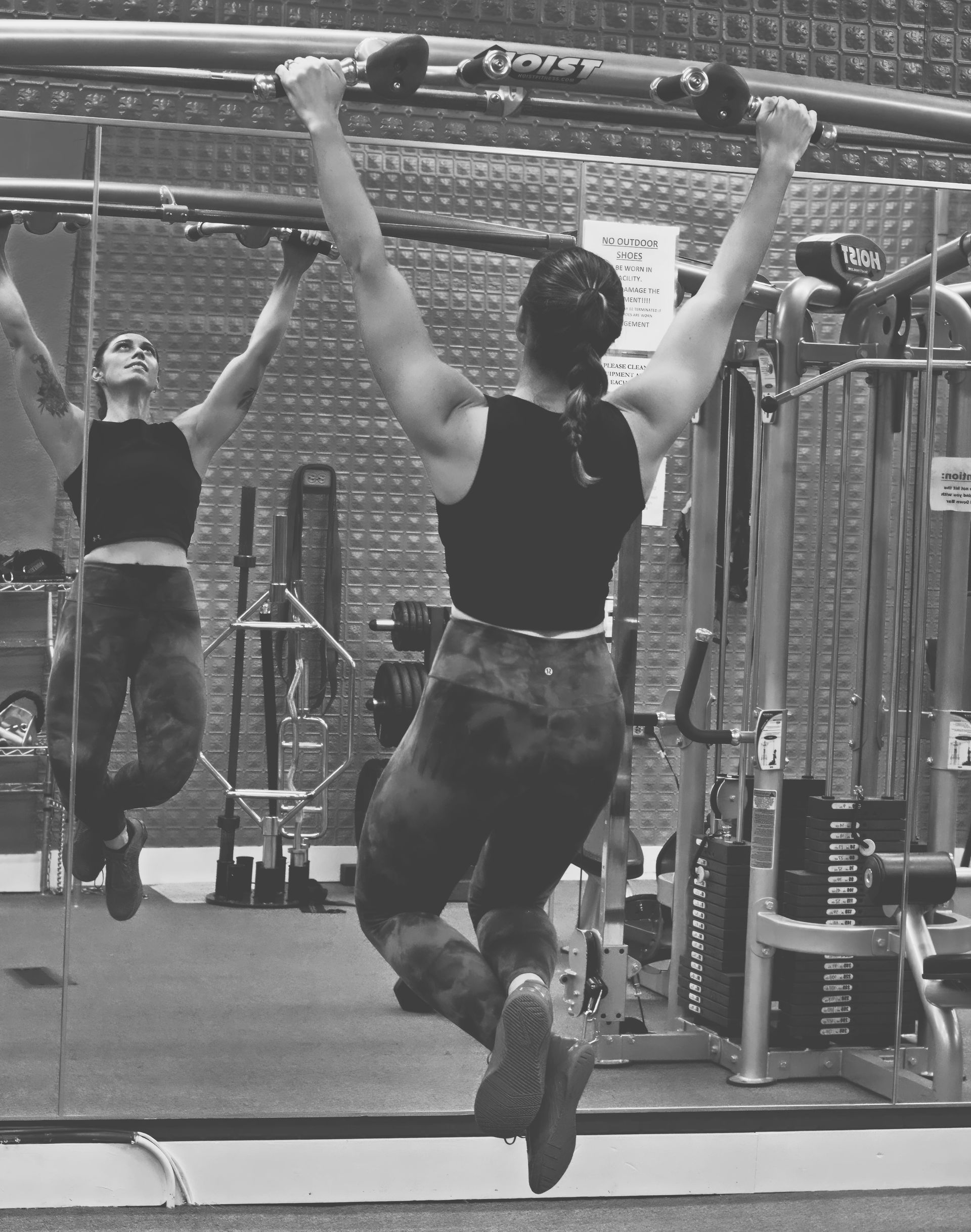 Person doing a pull-up in a gym, seen in a mirror, wearing a black tank top and camo pants.