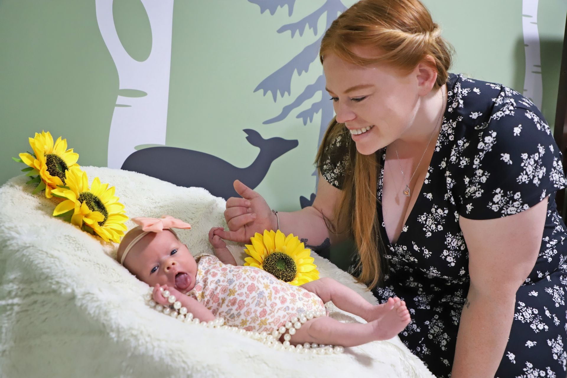 Woman smiling at a baby lying on a cushioned bed with sunflowers in a bright room