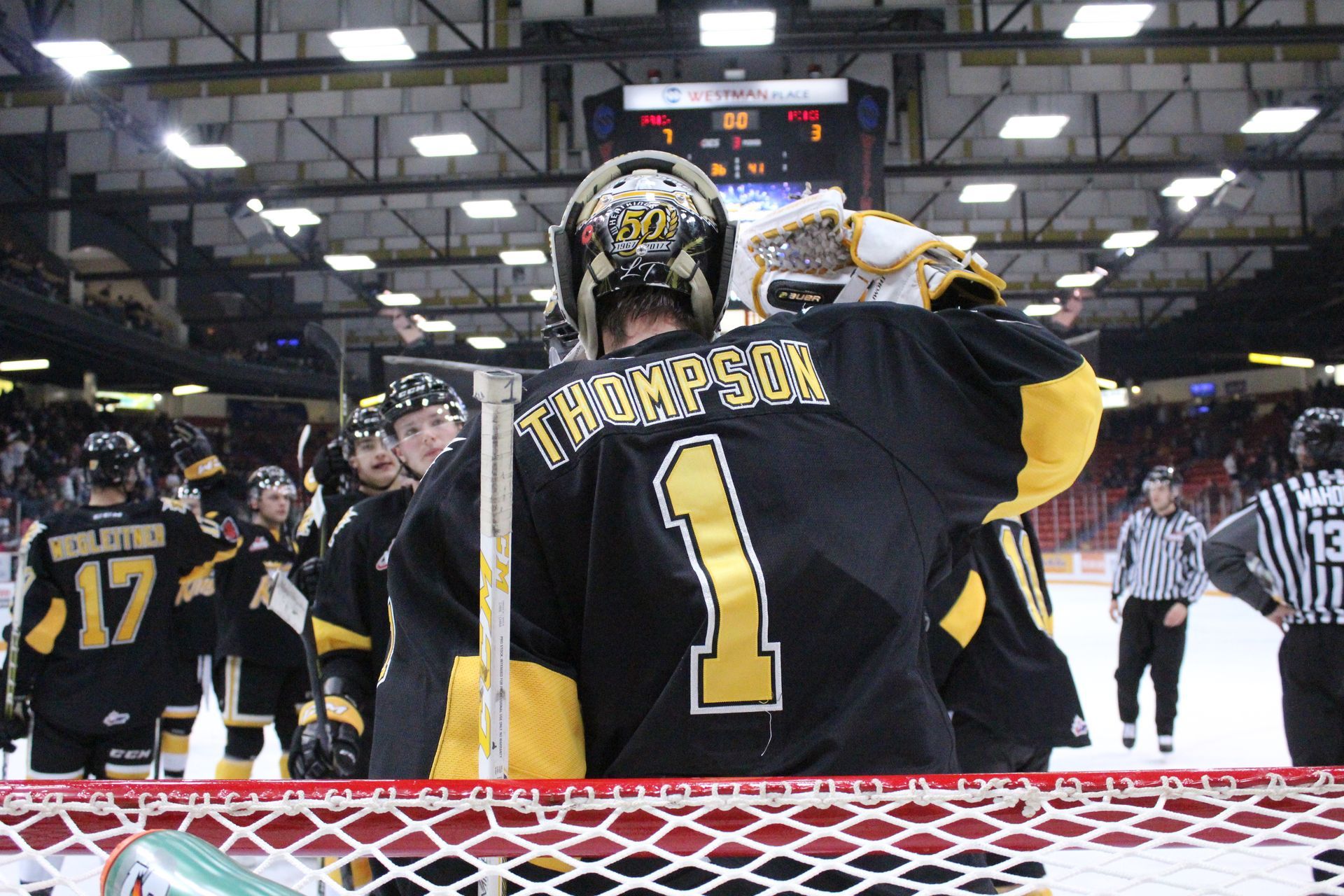 Hockey goalie in black and yellow jersey raising glove on rink after game, teammates nearby