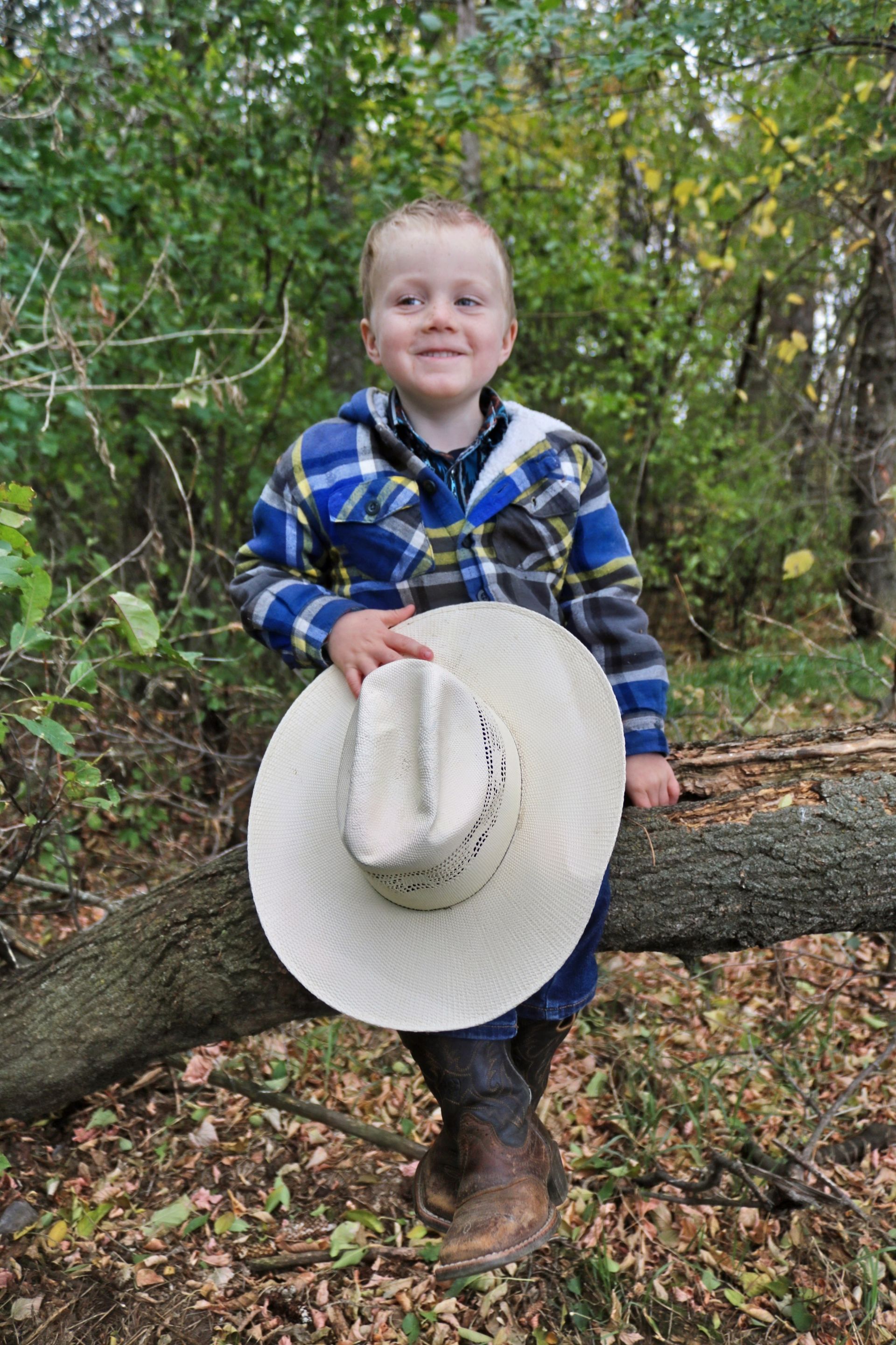 Child in a blue plaid shirt sitting on a tree branch, holding a white cowboy hat in a wooded area.