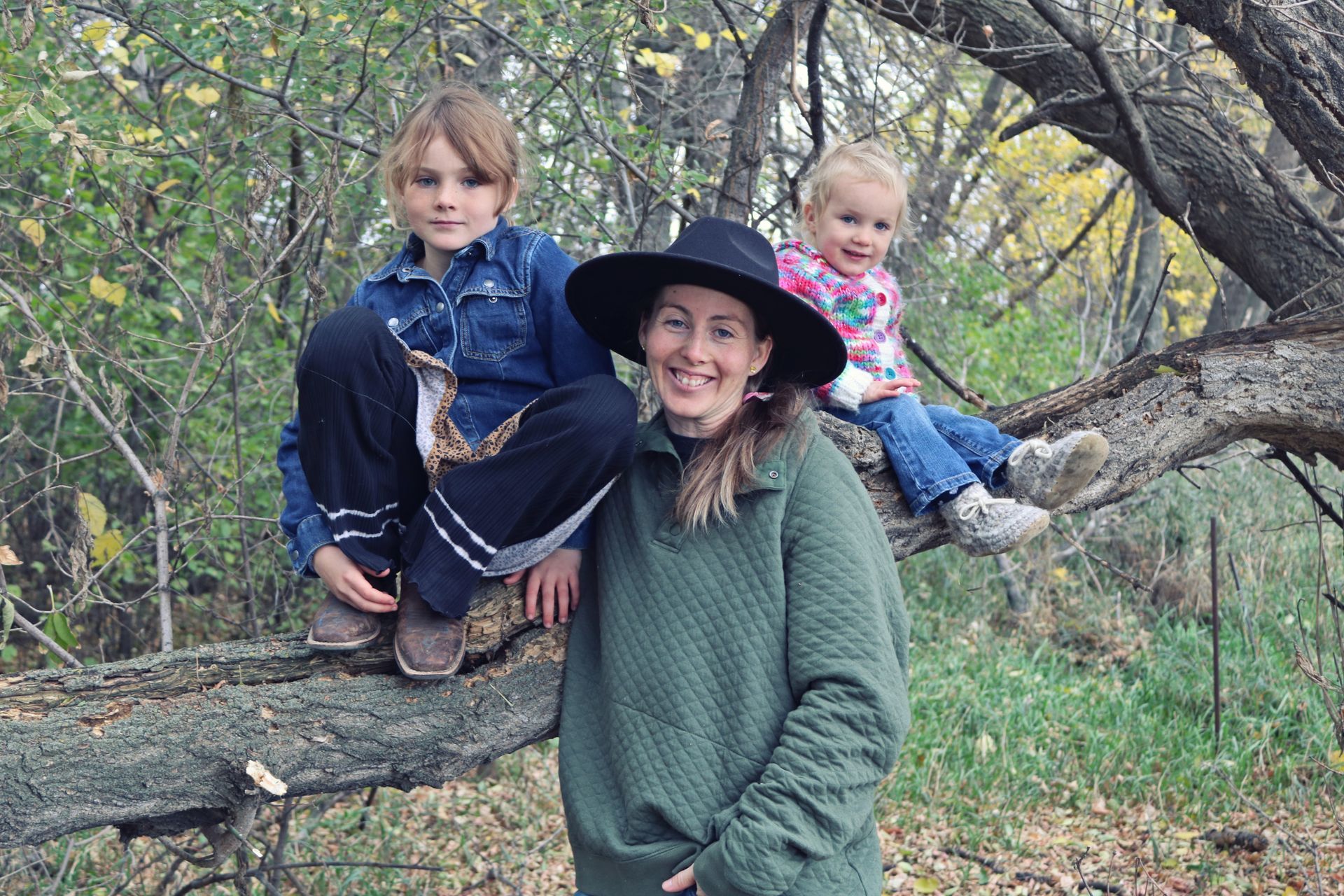 Three people sitting on a large tree branch in an autumn wooded area, smiling at the camera.