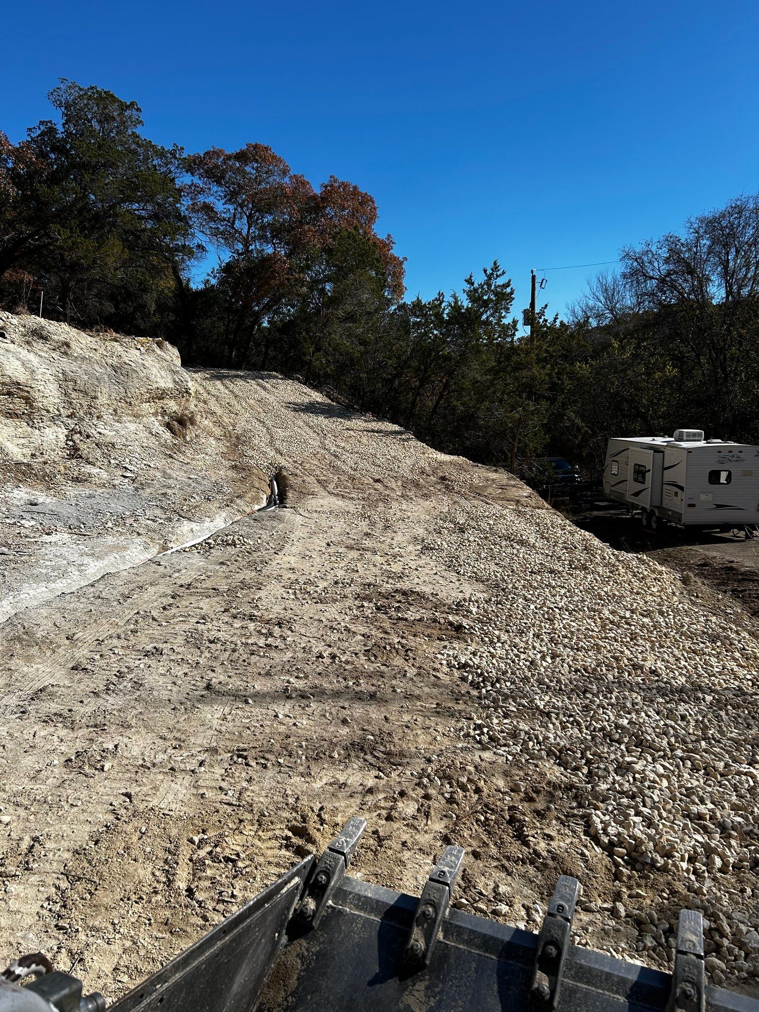 A pile of gravel beside a rocky hillside, under a blue sky, with trees in the background.