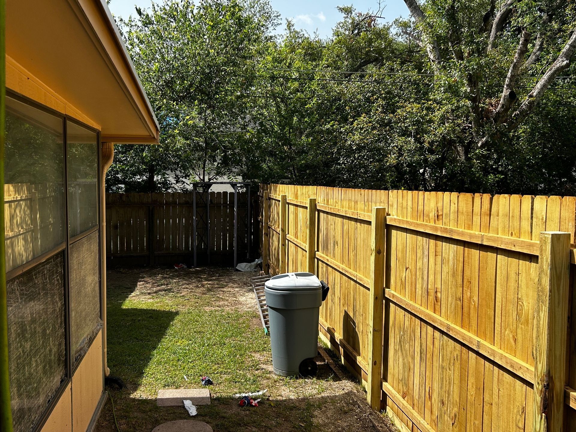 Backyard with wooden fence, trash can, and side of a yellow house.
