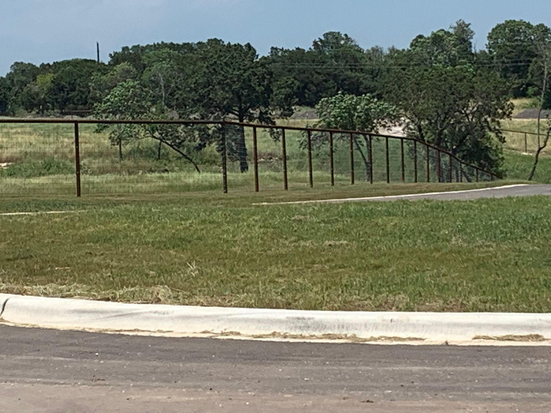 Chain-link fence borders a grassy field, trees in the background, a paved road in the foreground, cloudy sky.