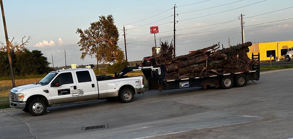 White truck pulling a trailer loaded with brown logs on a road near a yellow building.