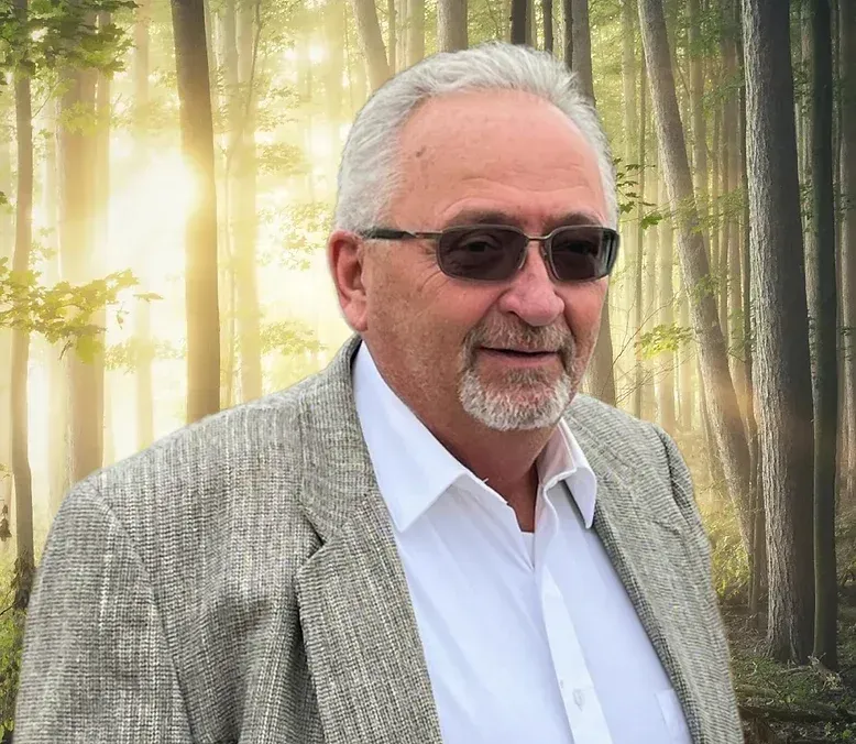Man in glasses and blazer smiles, set against a blurred forest backdrop.