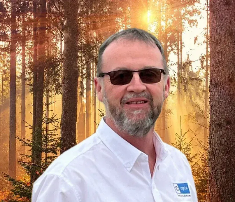 Man with a beard and sunglasses smiling, set against a forest background.