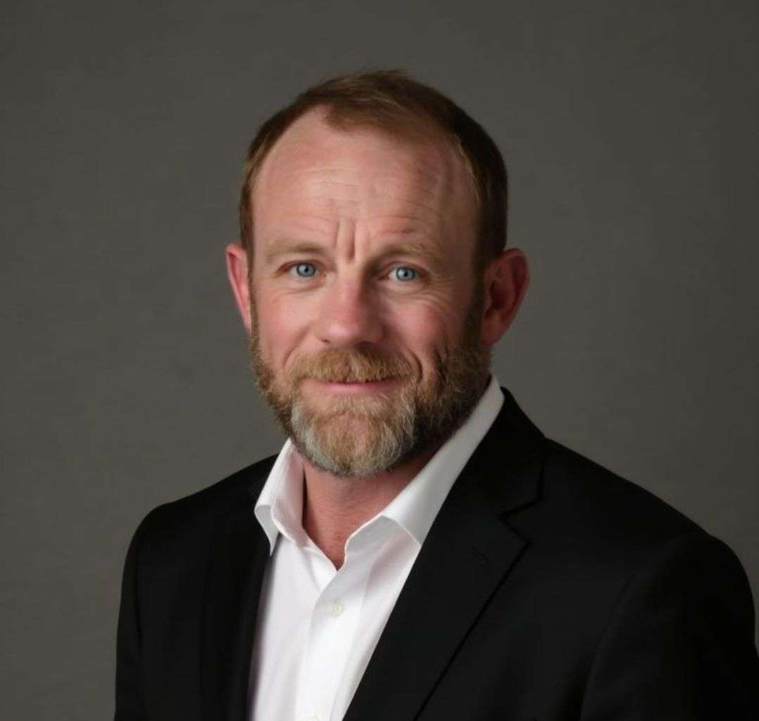 Man with a beard in a black suit and white shirt, looking directly at the camera with a neutral expression.