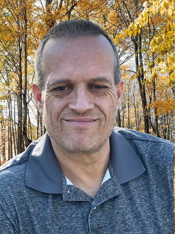 Man smiling, wearing a gray polo shirt, set against a blurred autumnal forest backdrop.