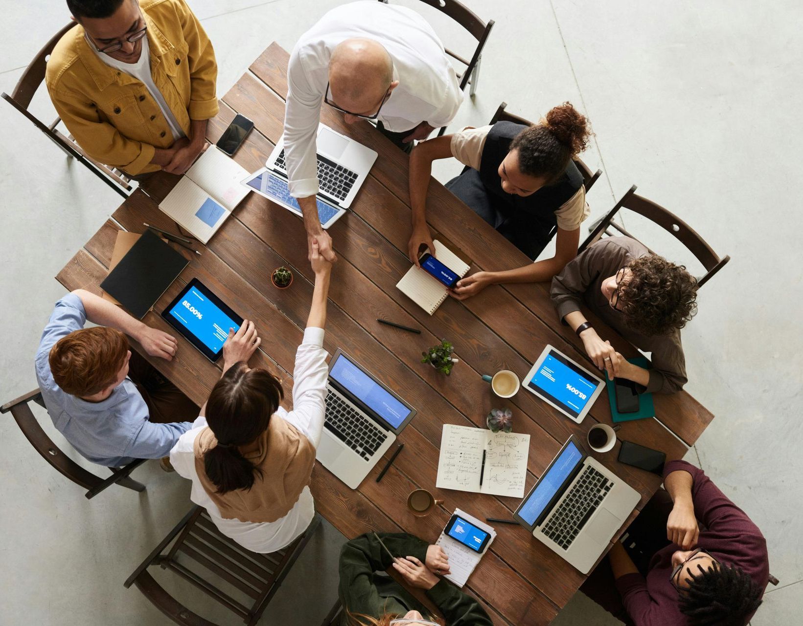 People at a wooden table in a meeting, two shaking hands, laptops and tablets visible.