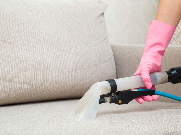 Person in pink gloves cleaning a beige sofa cushion with a cleaning tool.