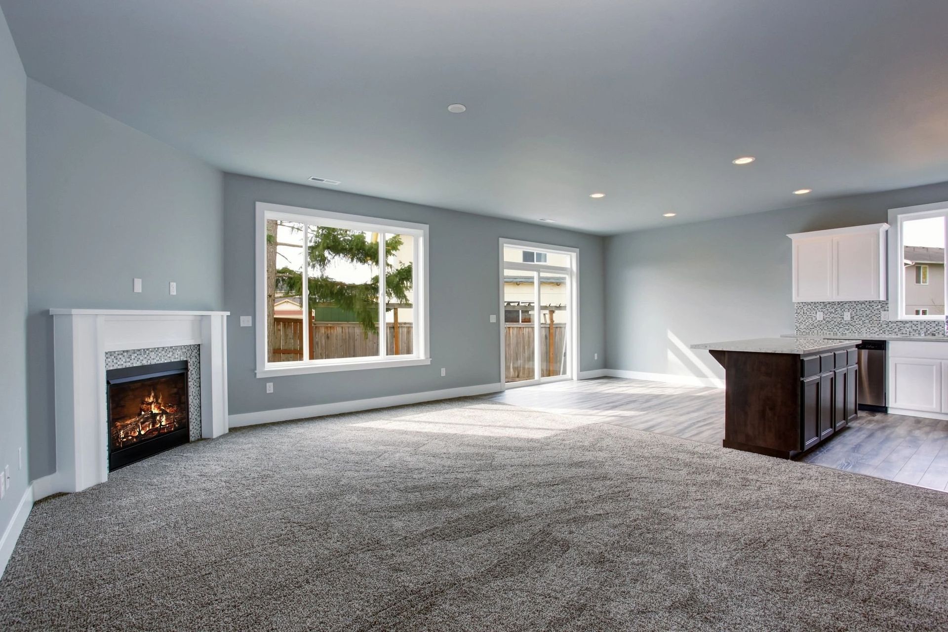 Spacious living room with gray walls, fireplace, large windows, dark brown island, and gray carpet.