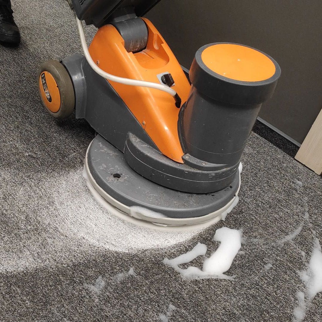 Orange and gray floor scrubber cleaning a carpet, creating foamy suds in an interior space.