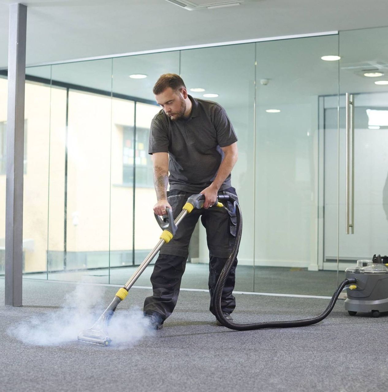 Man steam cleaning office carpet. He wears dark work clothes, steam visible from the cleaner.