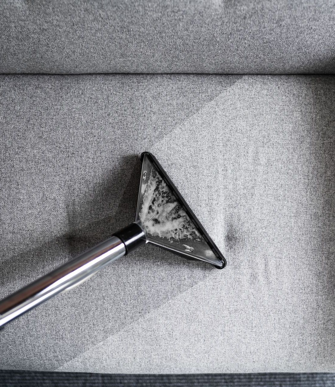 Vacuum nozzle cleaning gray upholstered sofa, removing dirt.
