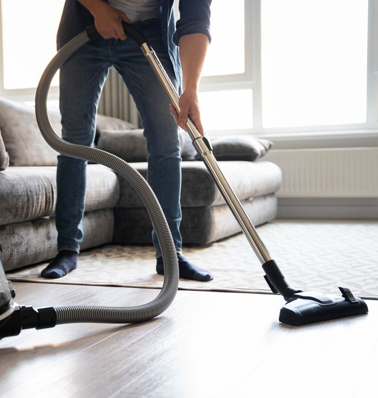 Person vacuuming a carpet in a living room, with a gray couch and hardwood floor.
