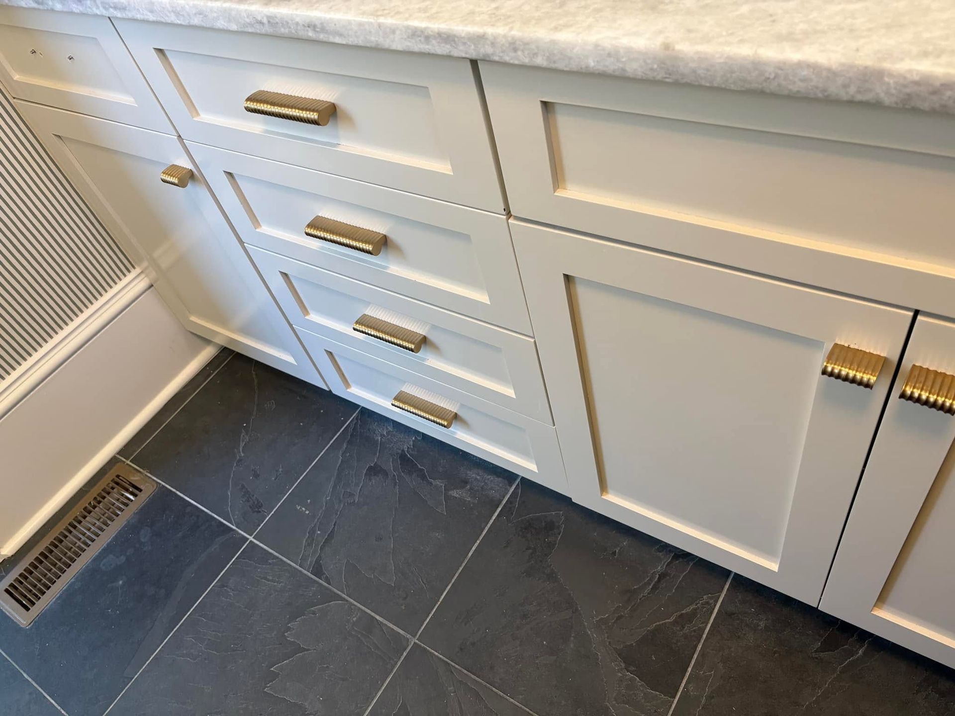 White kitchen cabinets with brass pulls and dark tile floor under a countertop.